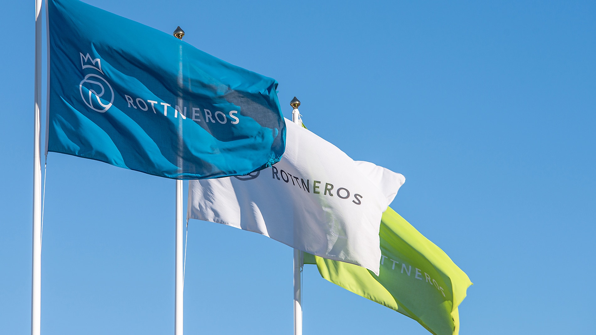 Tre flaggor som vajar mot en blå himmel. Flaggorna visar Rottneros logotyp och märke.