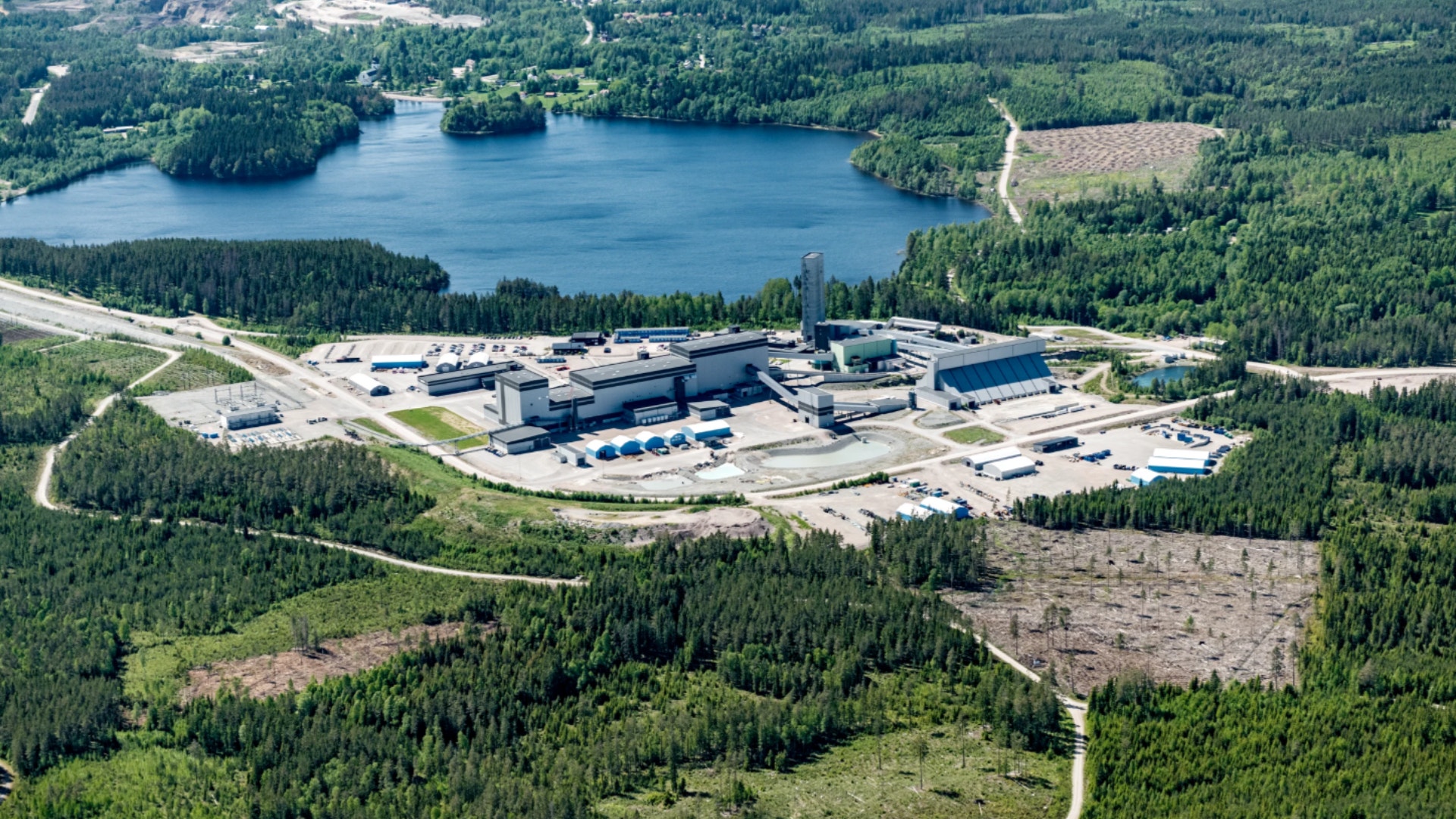 Flygfoto över en industrianläggning omgiven av skog och en sjö