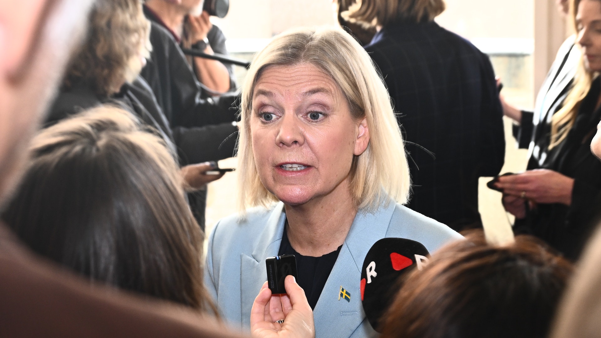 En ljushårig kvinna i ljusblå kavaj pratar med journalister som håller fram mikrofoner mot henne.