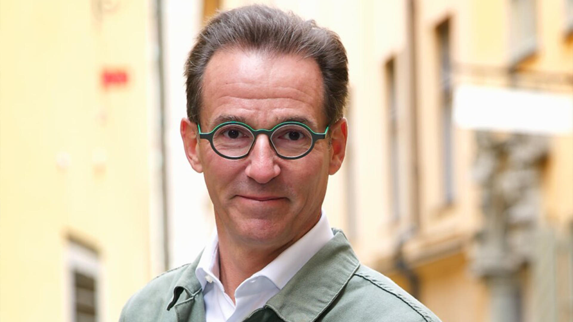 Porträtt av en man med glasögon och grå skjorta mot en suddig husfasad i bakgrunden.