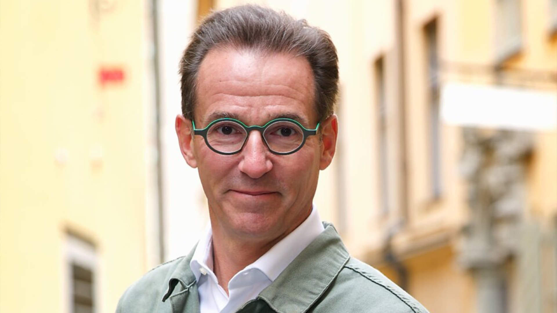 Porträtt av en man med glasögon och grå skjorta mot en suddig husfasad i bakgrunden.