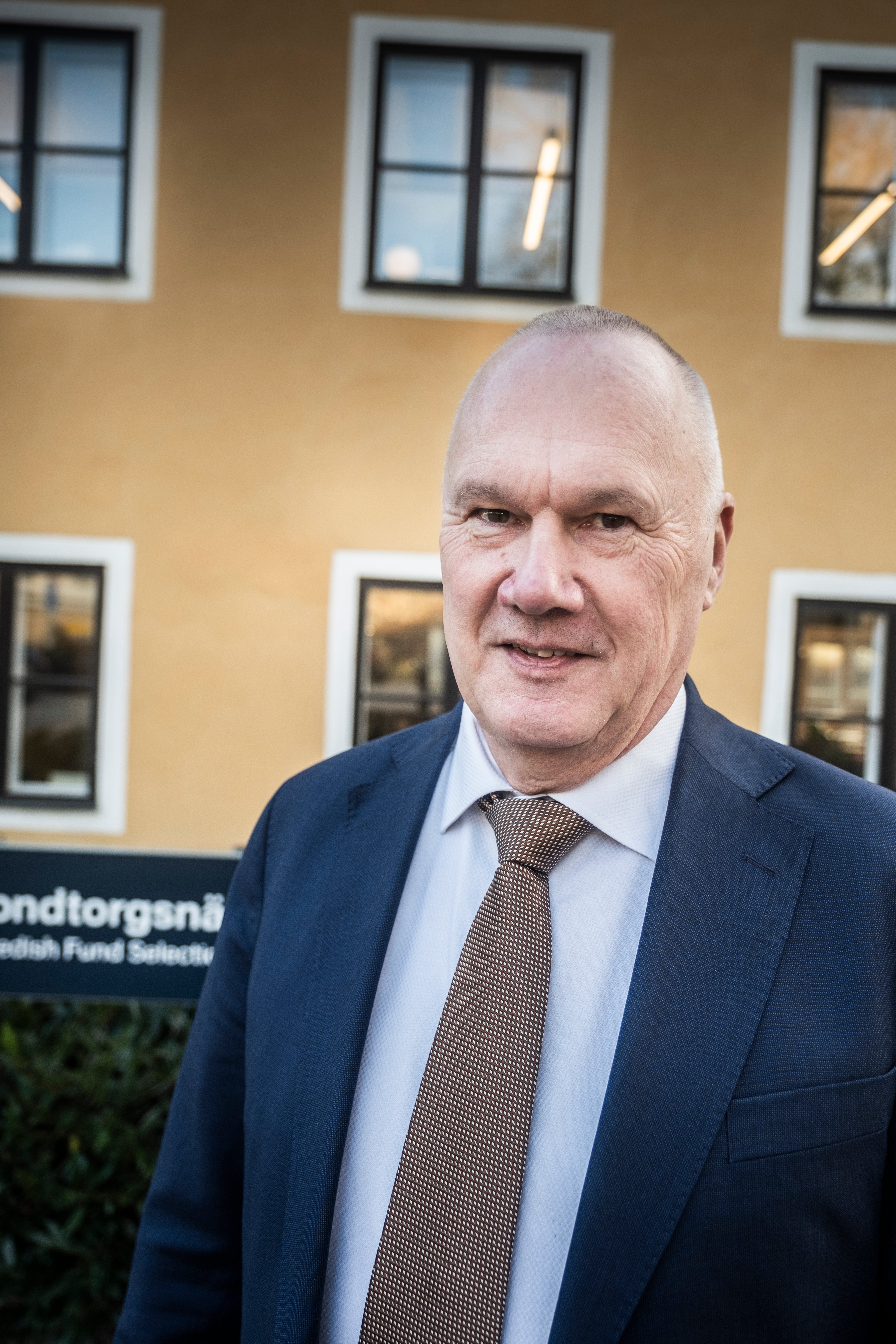 Porträtt av en man i mörkblå kostym och grå slips framför en gul byggnad med vita fönster.