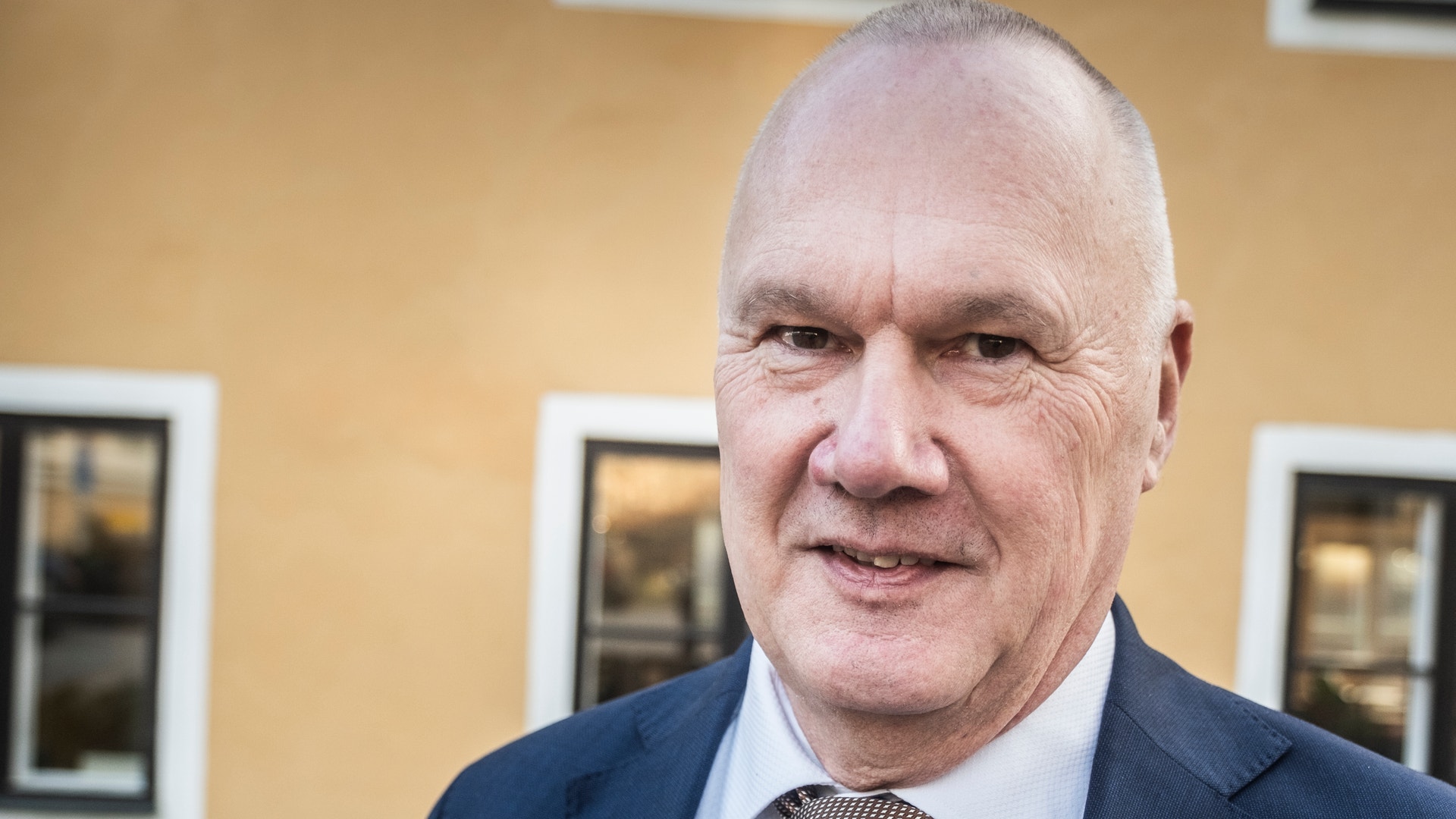 Porträtt av en man i mörkblå kostym och grå slips framför en gul byggnad med vita fönster.