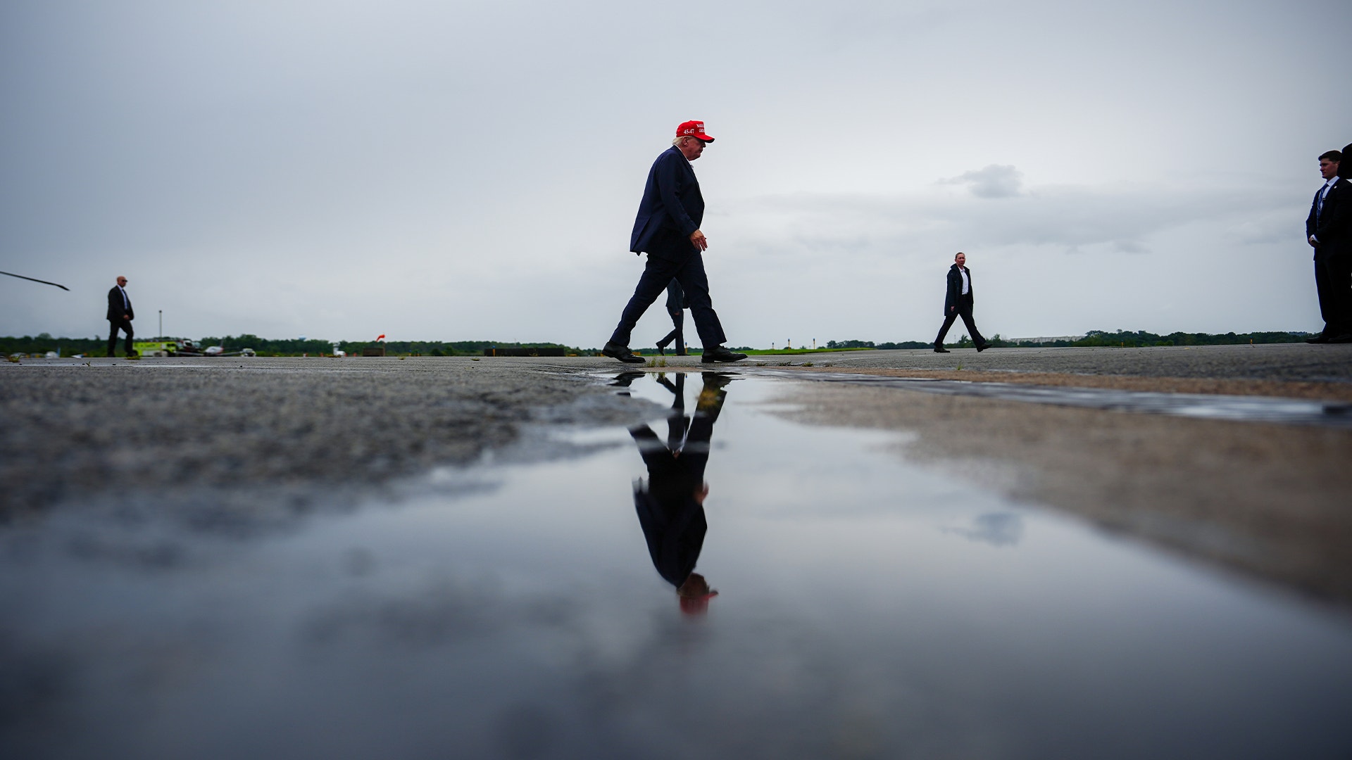Flera personer går på en våt, spegelblankt strand. En man i förgrunden bär röd mössa och mörk kostym. Hans reflektion syns tydligt i vattnet under honom. Molnig himmel i bakgrunden.