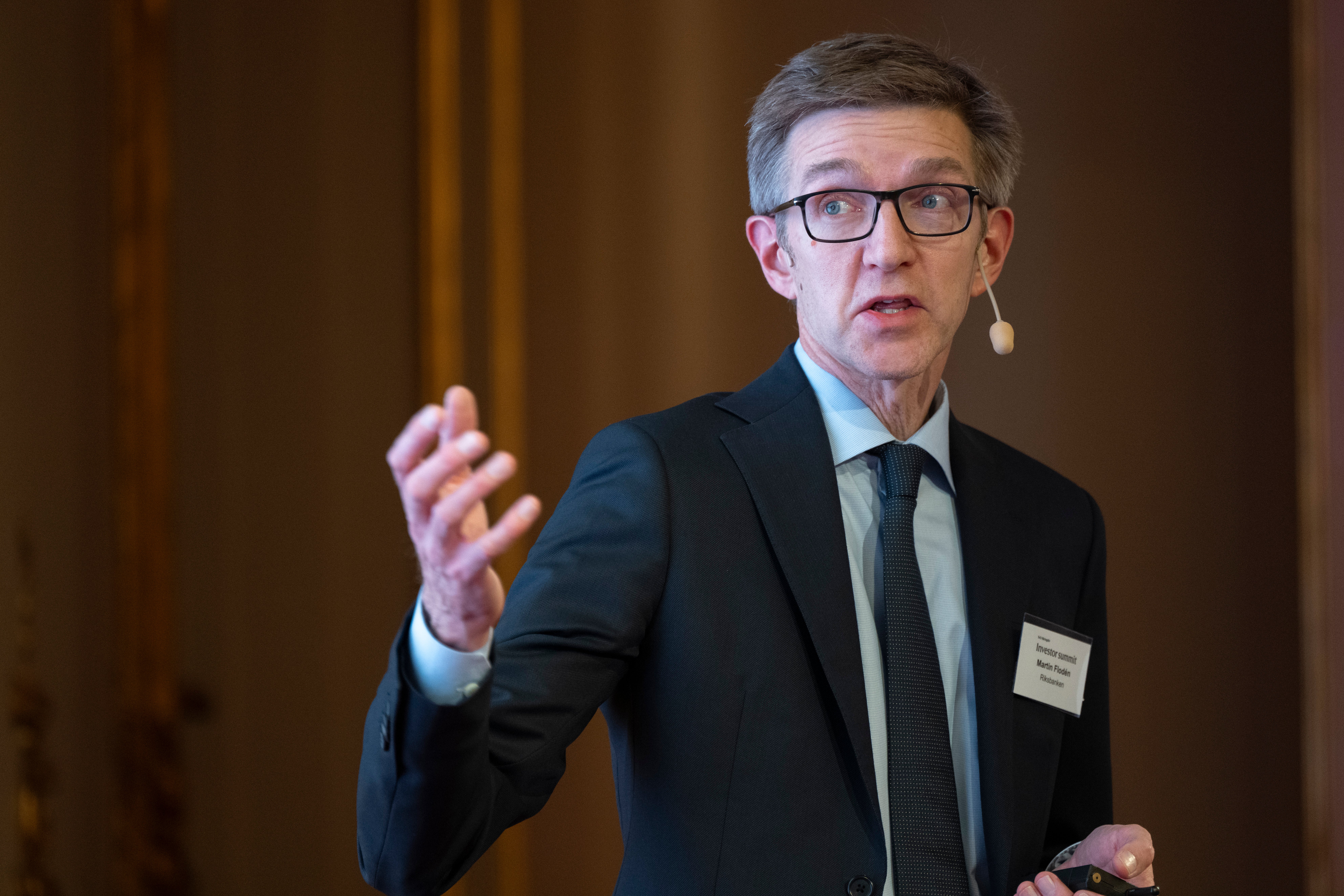 Man i mörk kostym och glasögon håller en presentation och gestikulerar med handen.