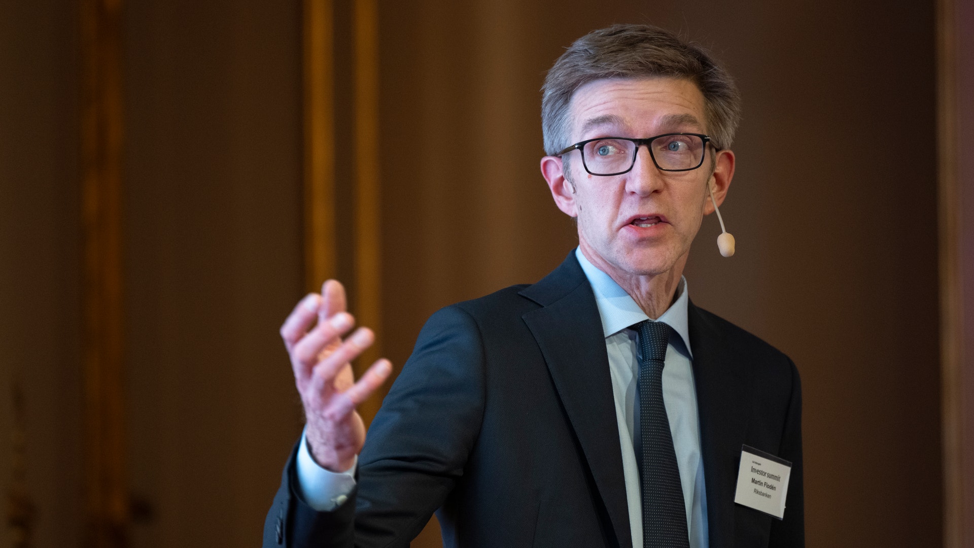 Man i mörk kostym och glasögon håller en presentation och gestikulerar med handen.