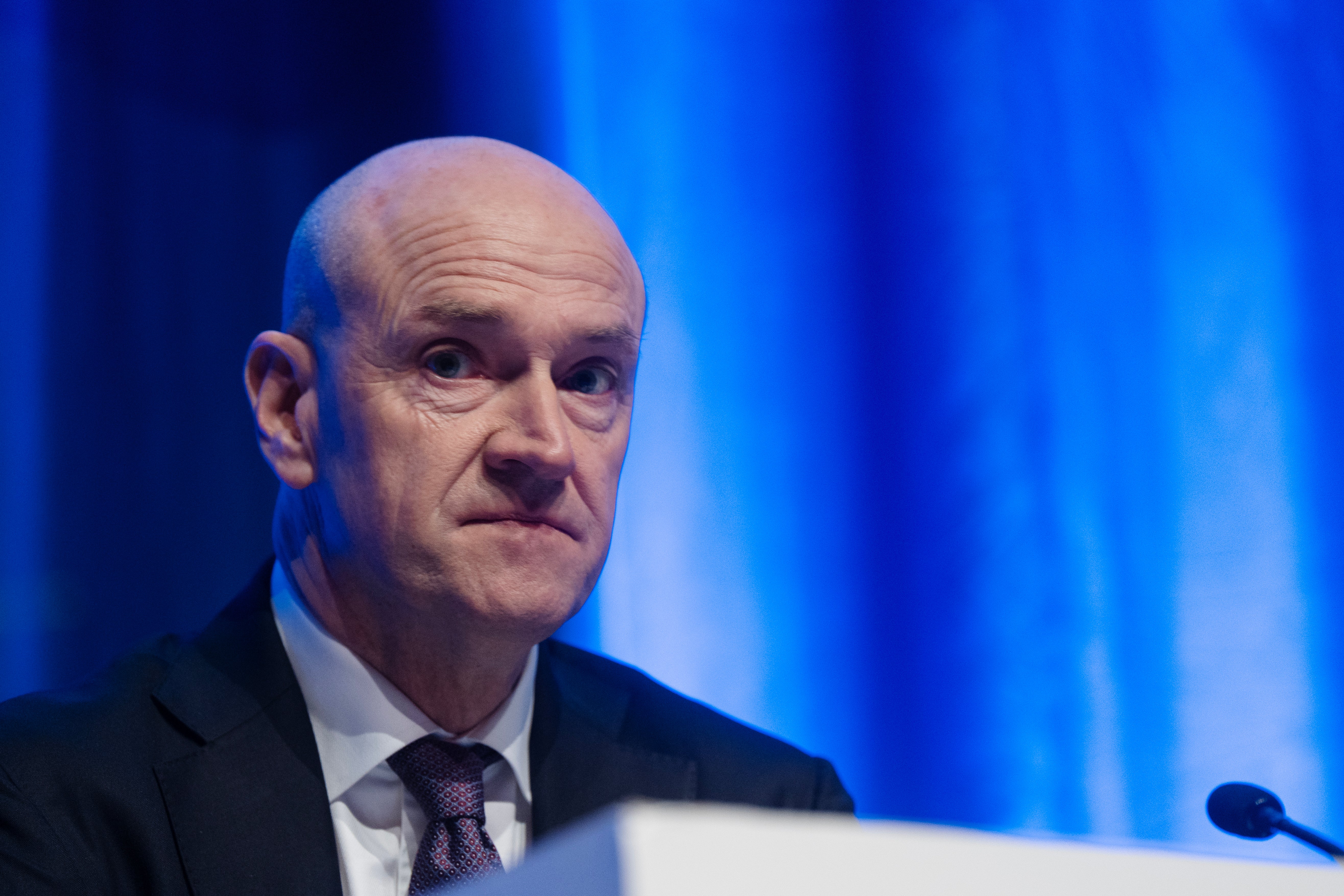 En man i kostym talar vid en podium framför en blå bakgrund.