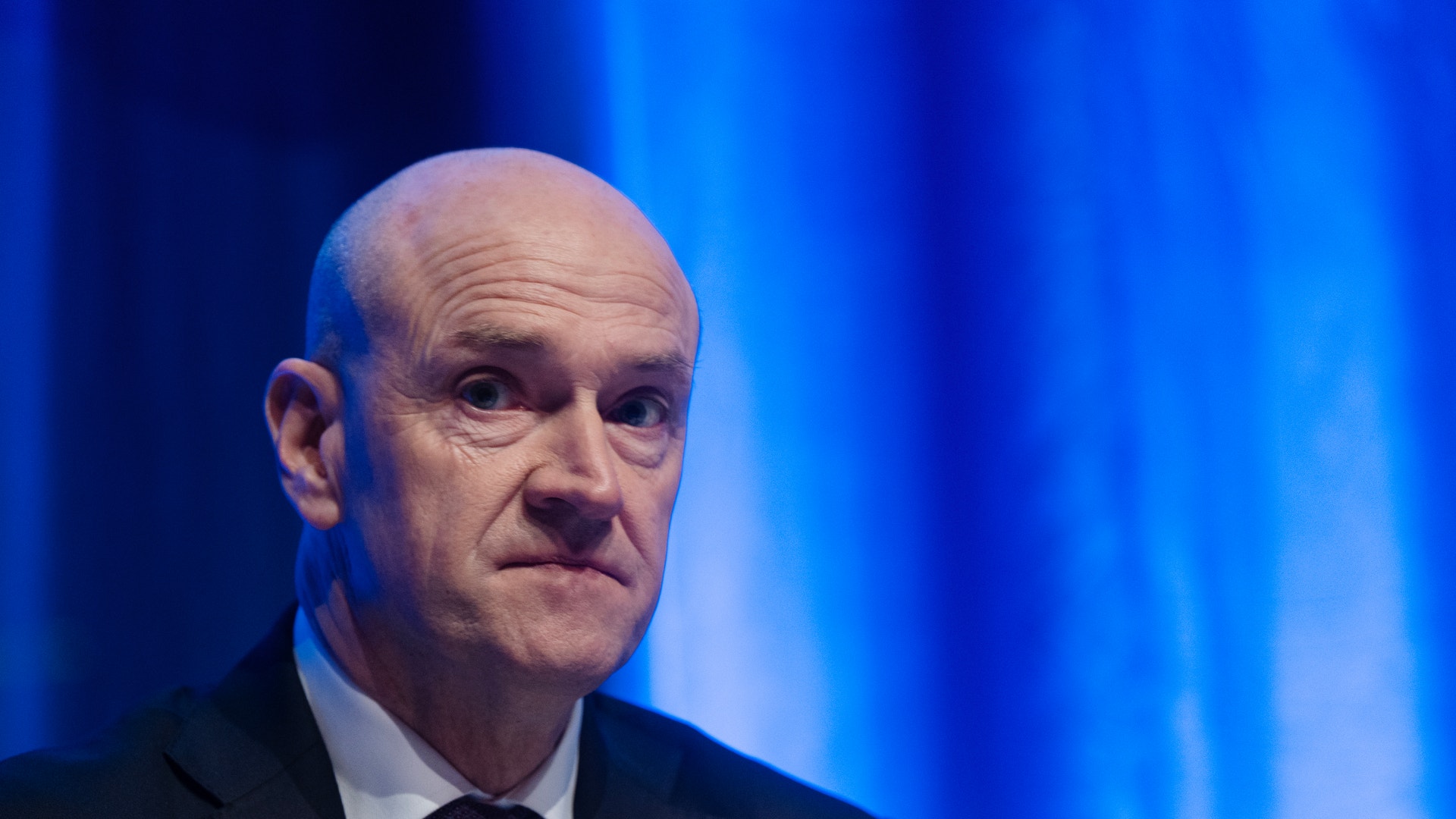 En man i kostym talar vid en podium framför en blå bakgrund.