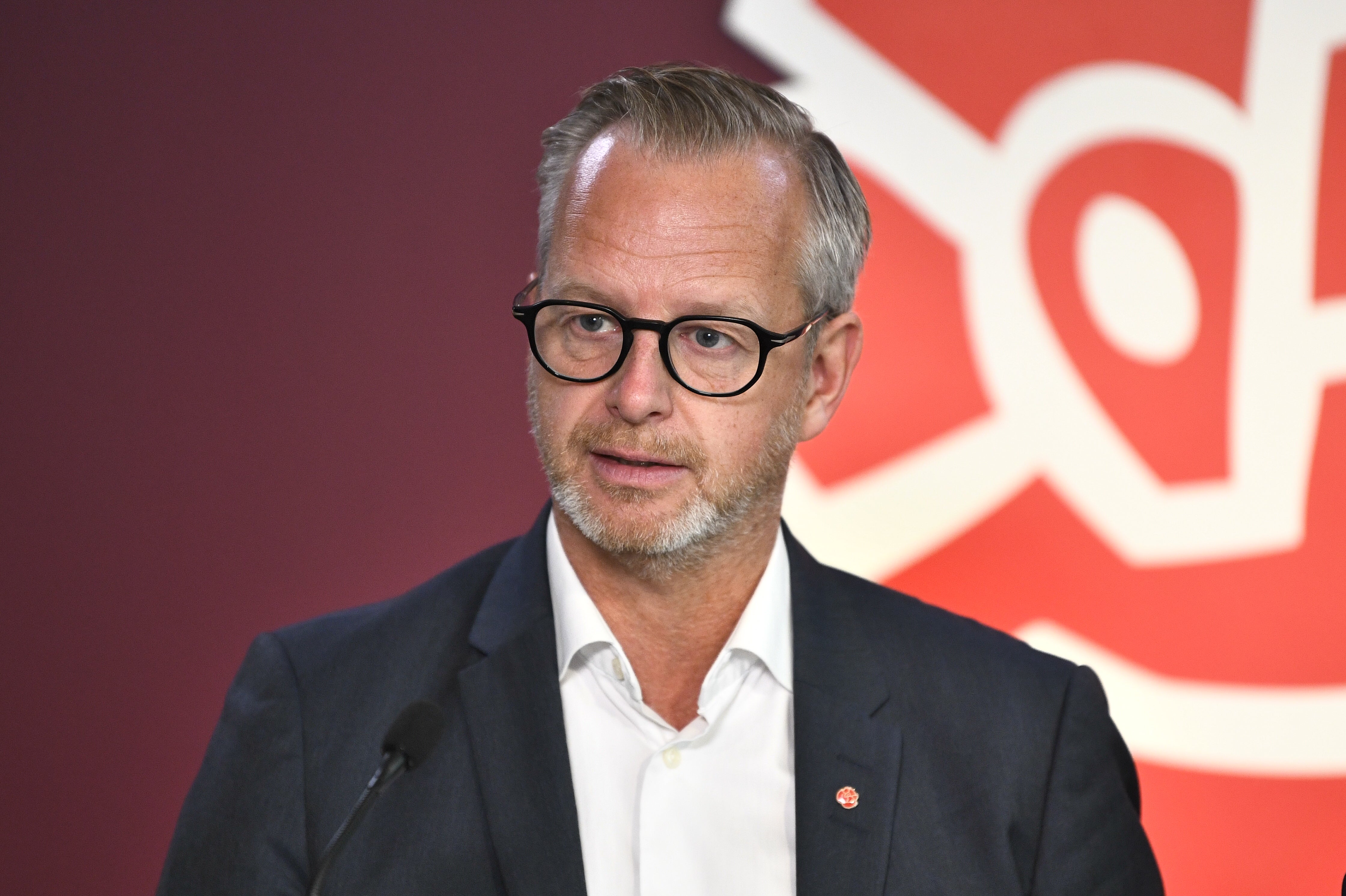 En man med glasögon och mörkblå kavaj framför en röd bakgrund.