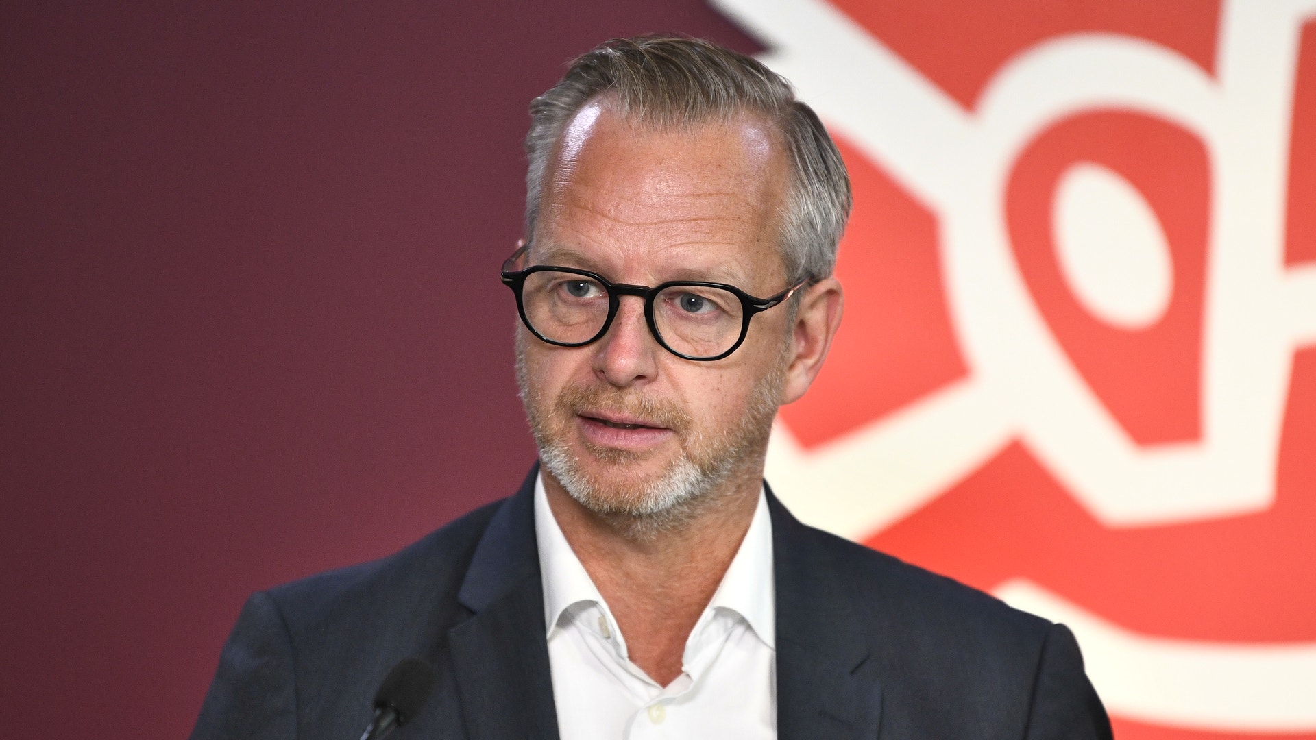 En man med glasögon och mörkblå kavaj framför en röd bakgrund.