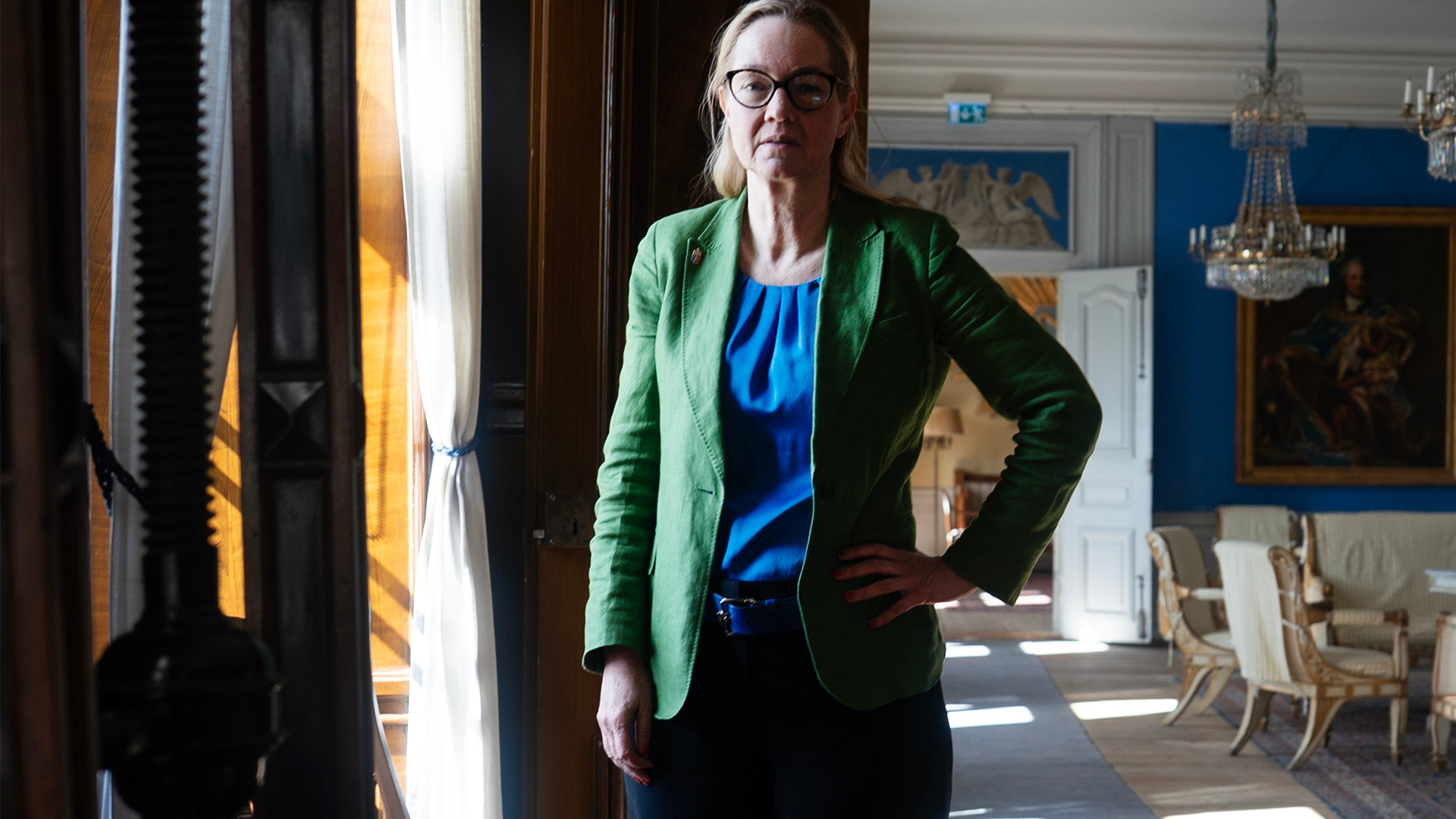 Porträtt av en person med glasögon, grön blazer och blå blus som står i ett klassiskt inredningsrum med högt i tak och elegant möblering.