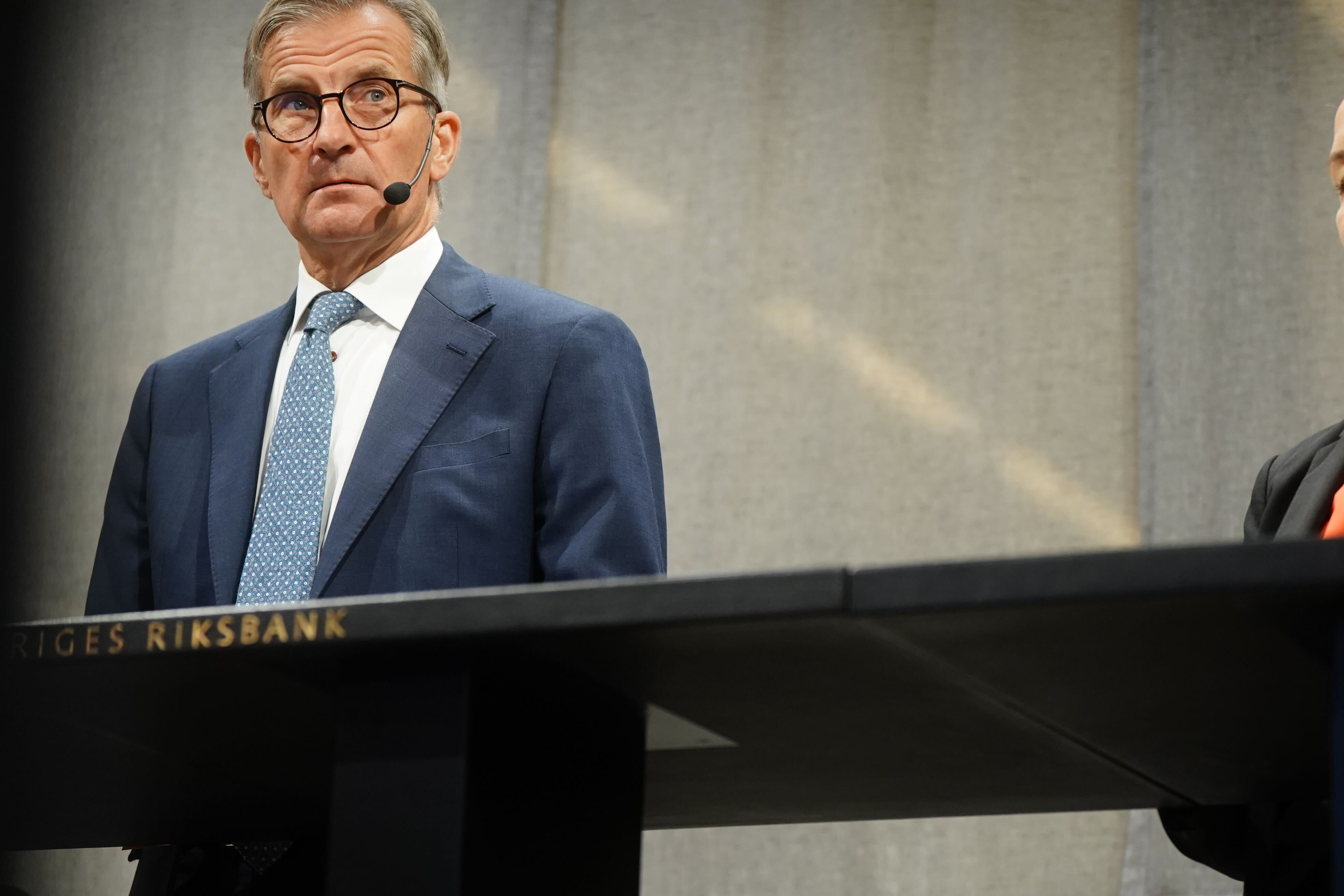 Man i blå kostym och glasögon står vid ett podium märkt Sveriges Riksbank och håller ett tal.