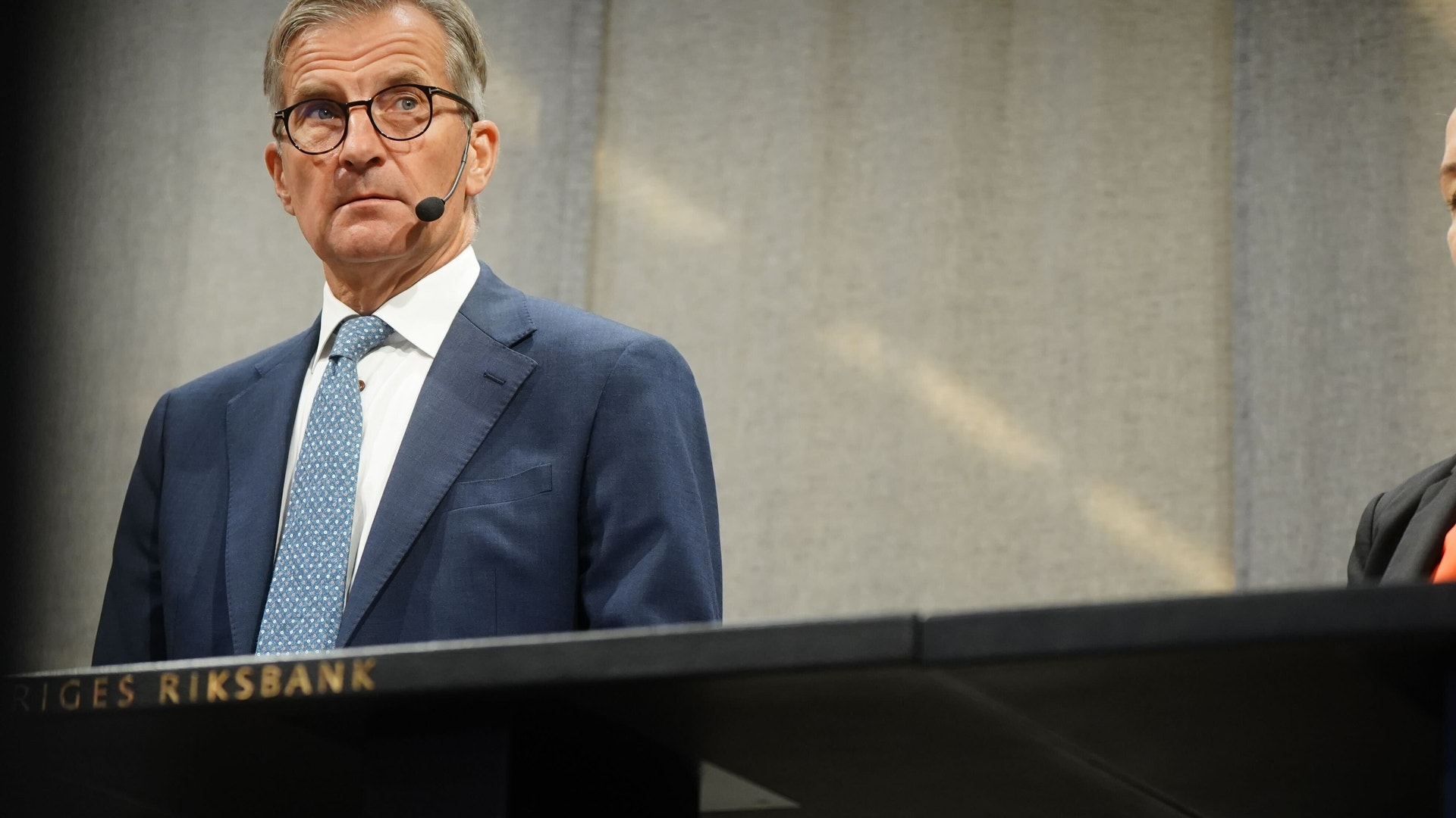 Man i blå kostym och glasögon står vid ett podium märkt Sveriges Riksbank och håller ett tal.