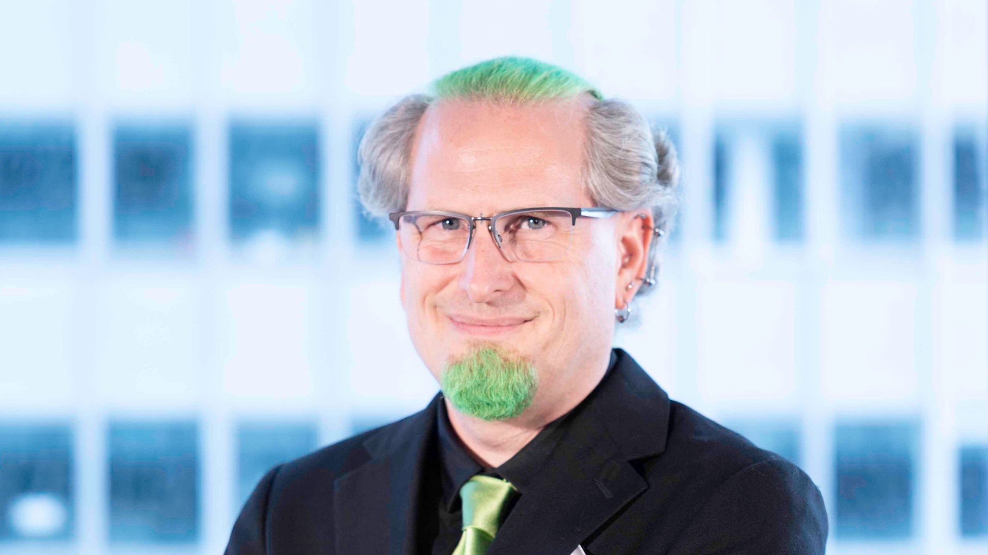 Porträtt av en leende man med glasögon, grått hår och grönfärgad hårtull och haka, klädd i svart kavaj och grön slips.