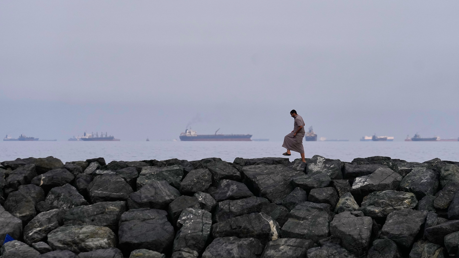 Person står på en stenig bränning med fartyg synliga på havet i bakgrunden