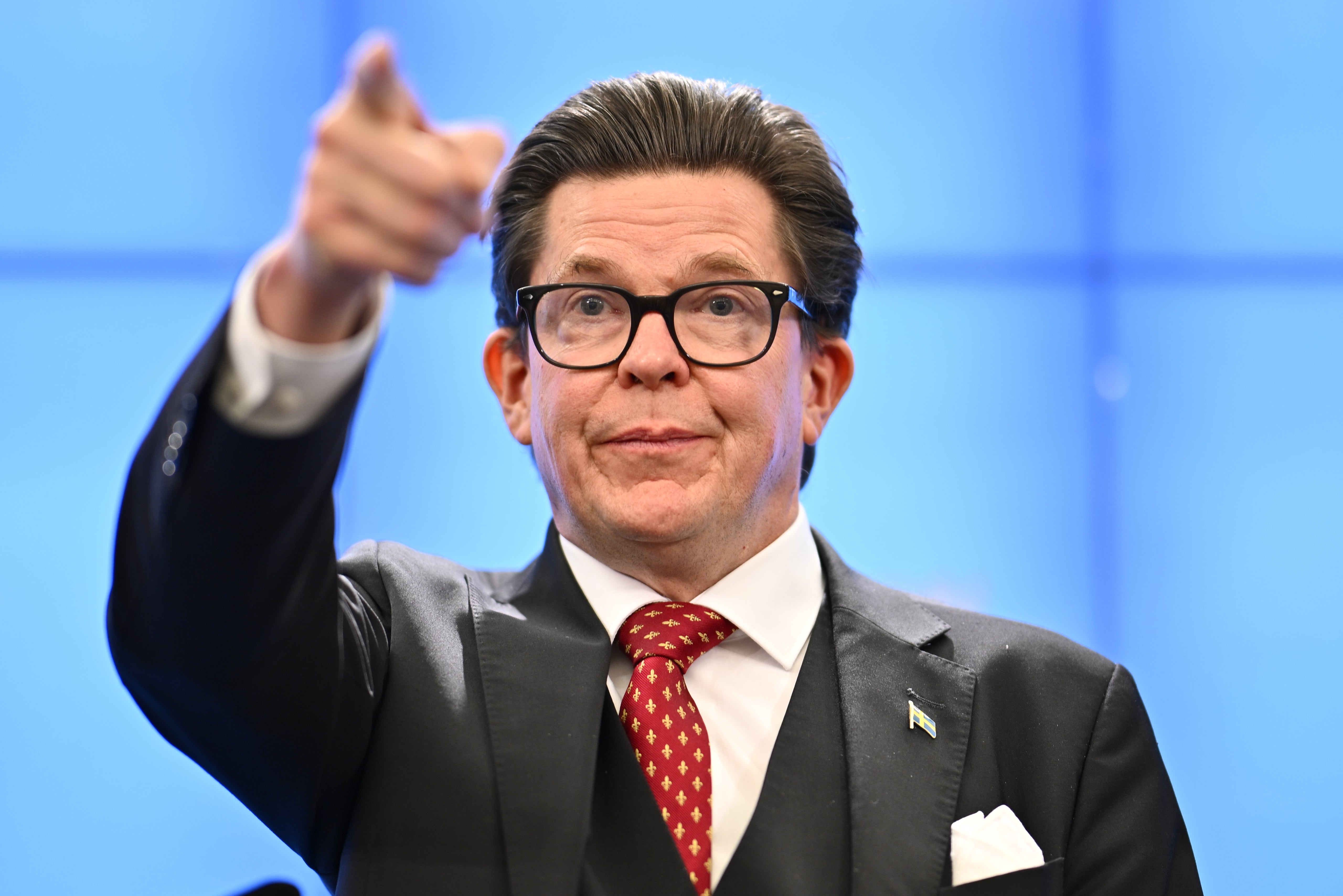 Andreas Norlén i mörk kostym och glasögon pekar mot kameran framför en blå bakgrund.