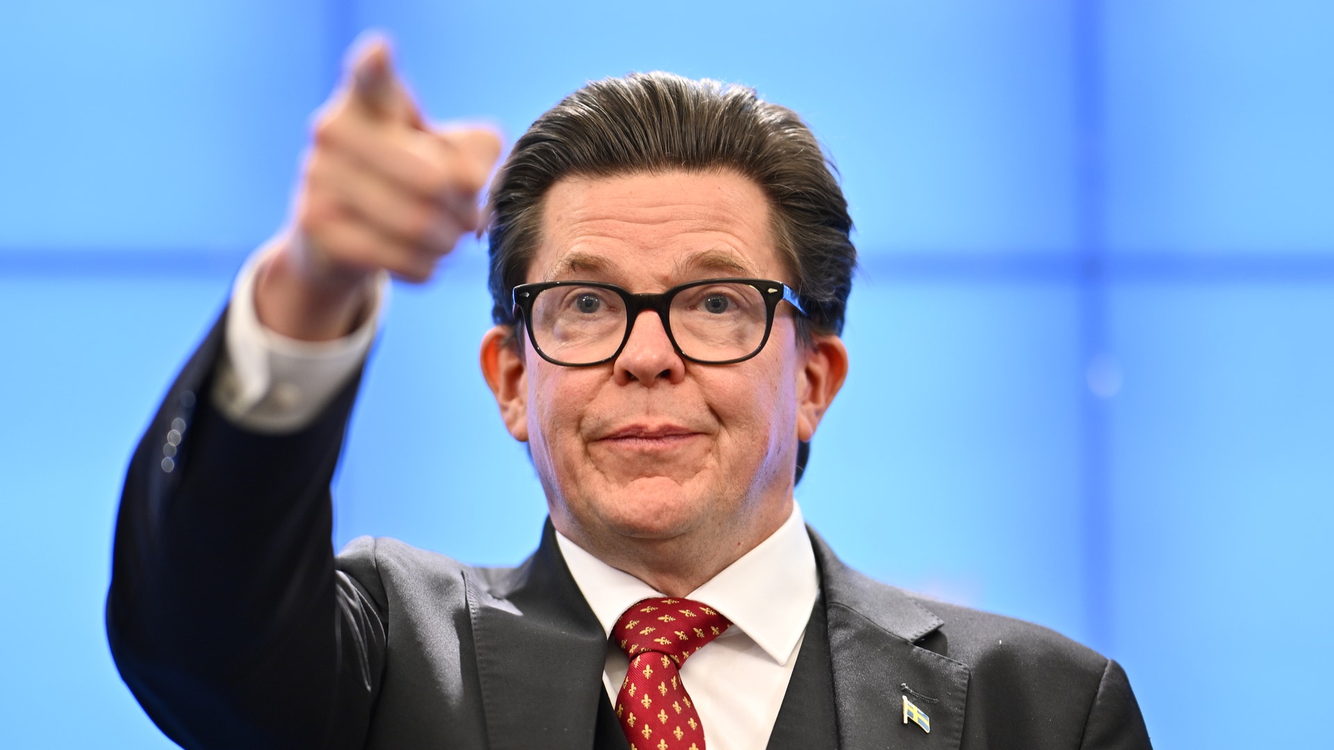 Andreas Norlén i mörk kostym och glasögon pekar mot kameran framför en blå bakgrund.