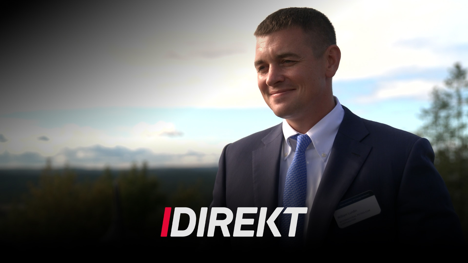 En man i kostym och slips ler utomhus, med en skog och himmel i bakgrunden. Texten "DIREKT" syns i bild.