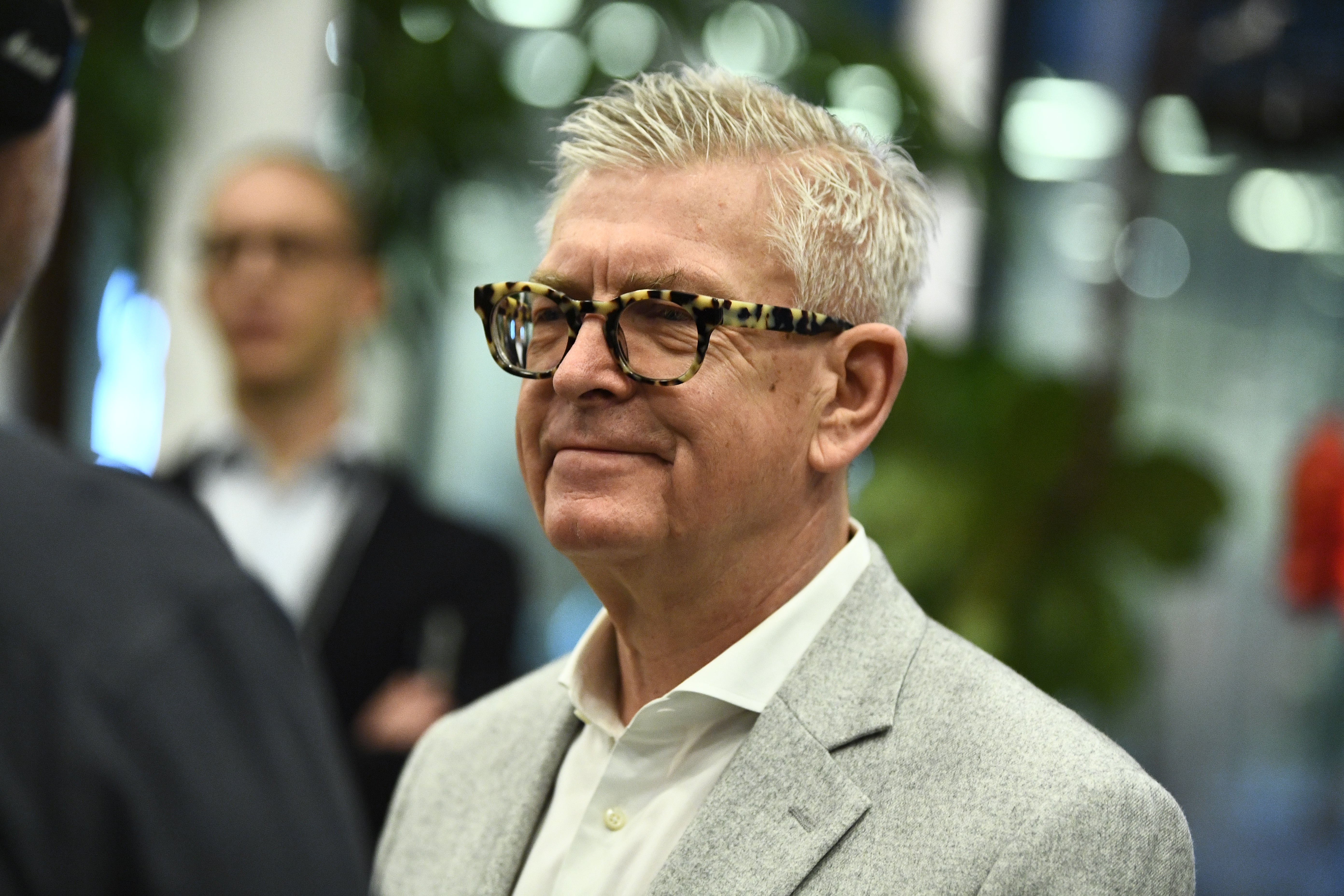 En äldre man med vitt hår och glasögon, klädd i ljusgrå kavaj och vit skjorta, fotograferad utomhus.