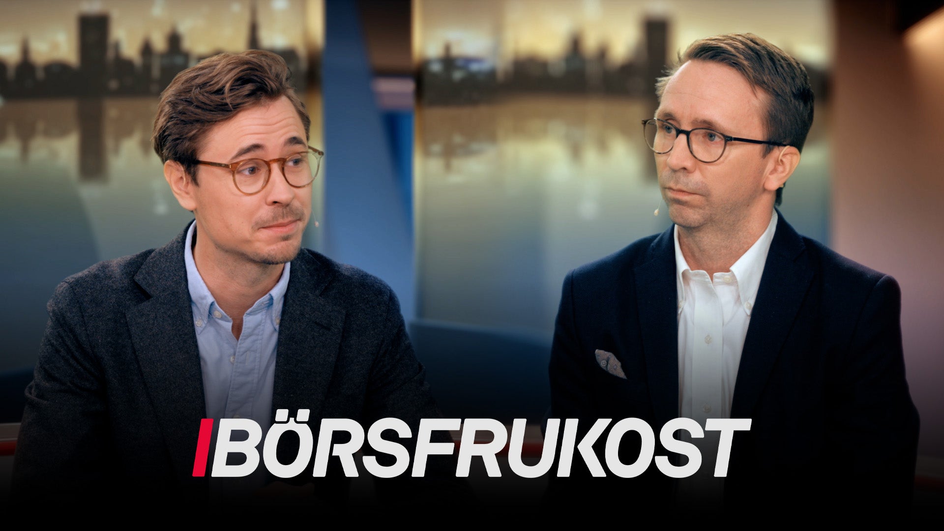 Två män i affärskläder diskuterar i en tv-studio med stadsvy i bakgrunden