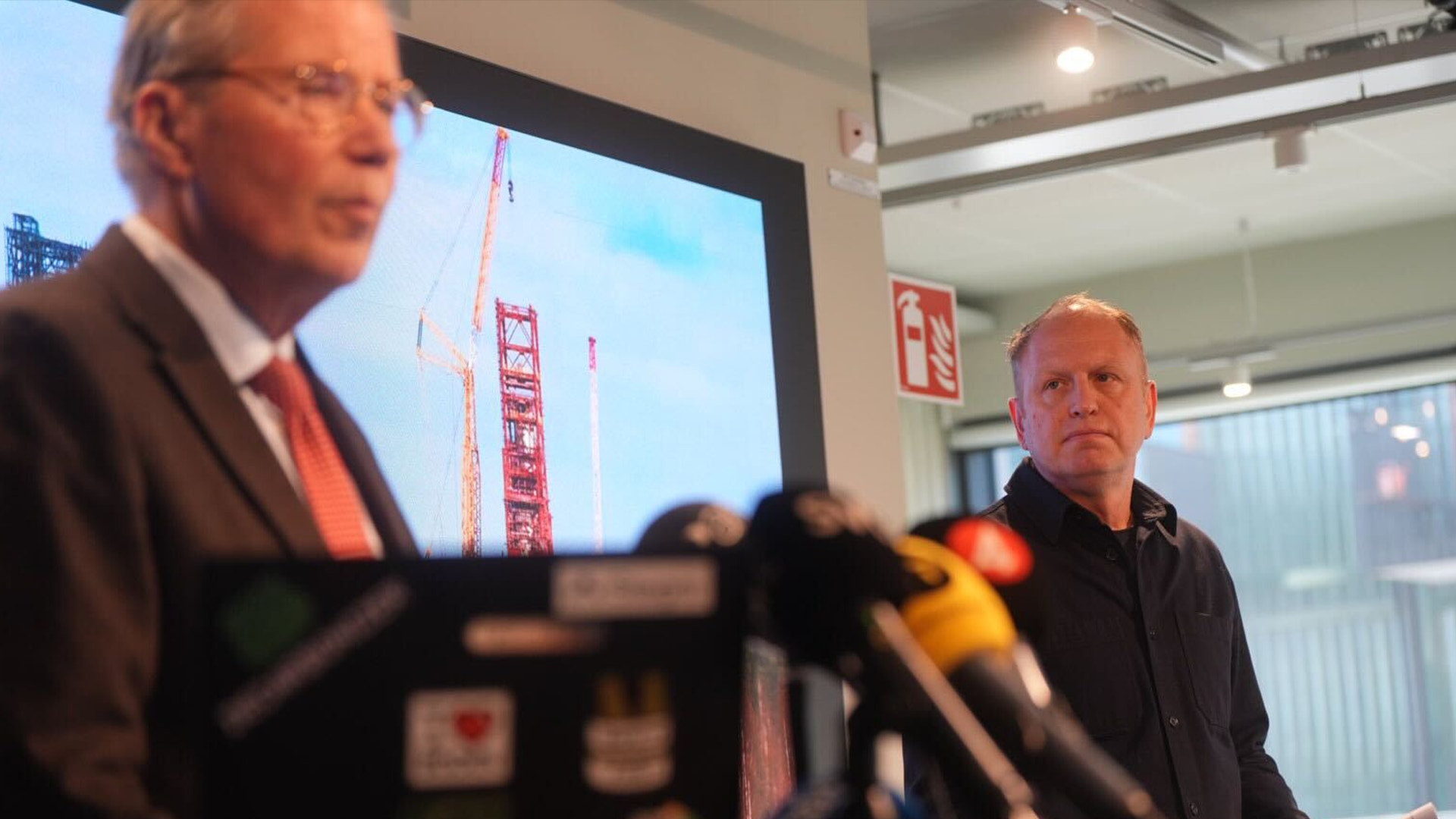 Två män vid en presskonferens med en skärm som visar en byggkran i bakgrunden.