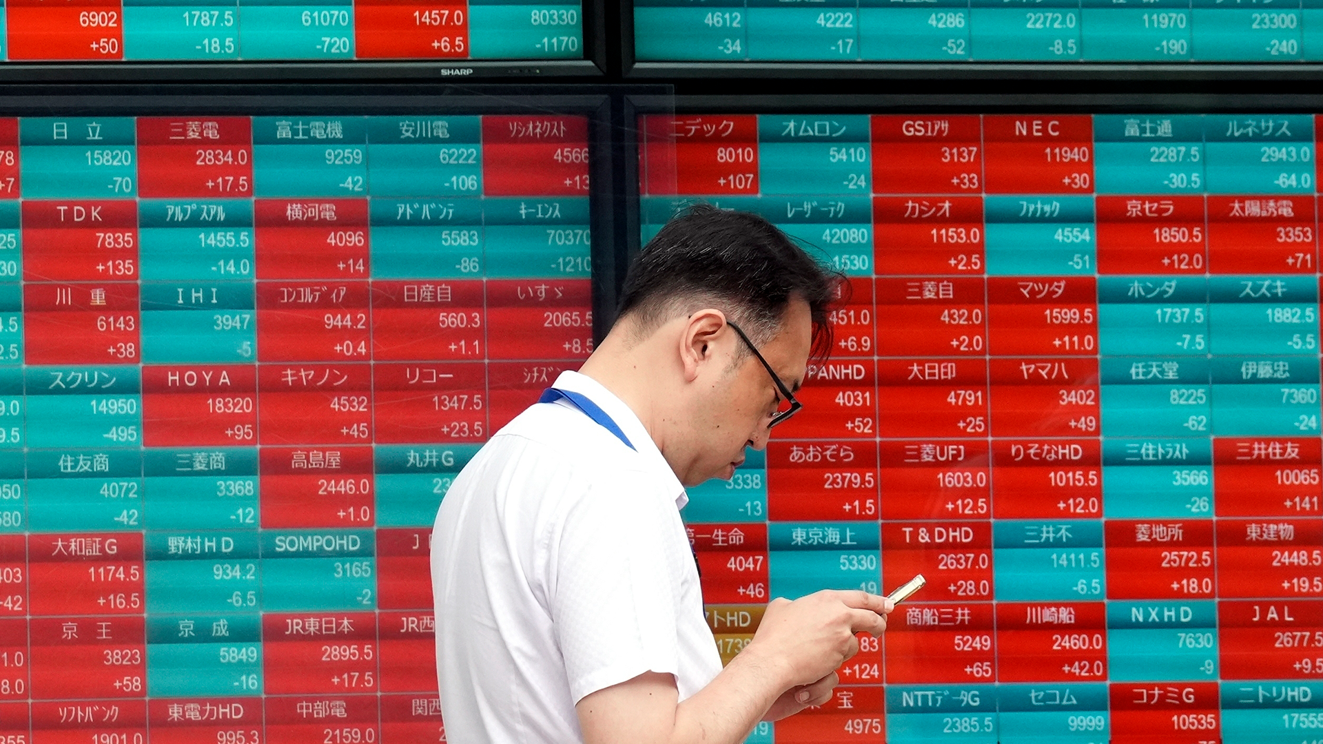 Man i vit skjorta och glasögon står framför en stor elektronisk skärm med finansiell data och japanska tecken.