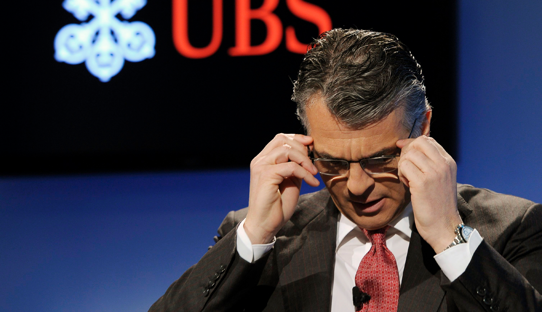 Man i mörk kostym och röd slips justerar sina glasögon på en scen med UBS-logotyp i bakgrunden.Sergio Ermotti, chef UBS.