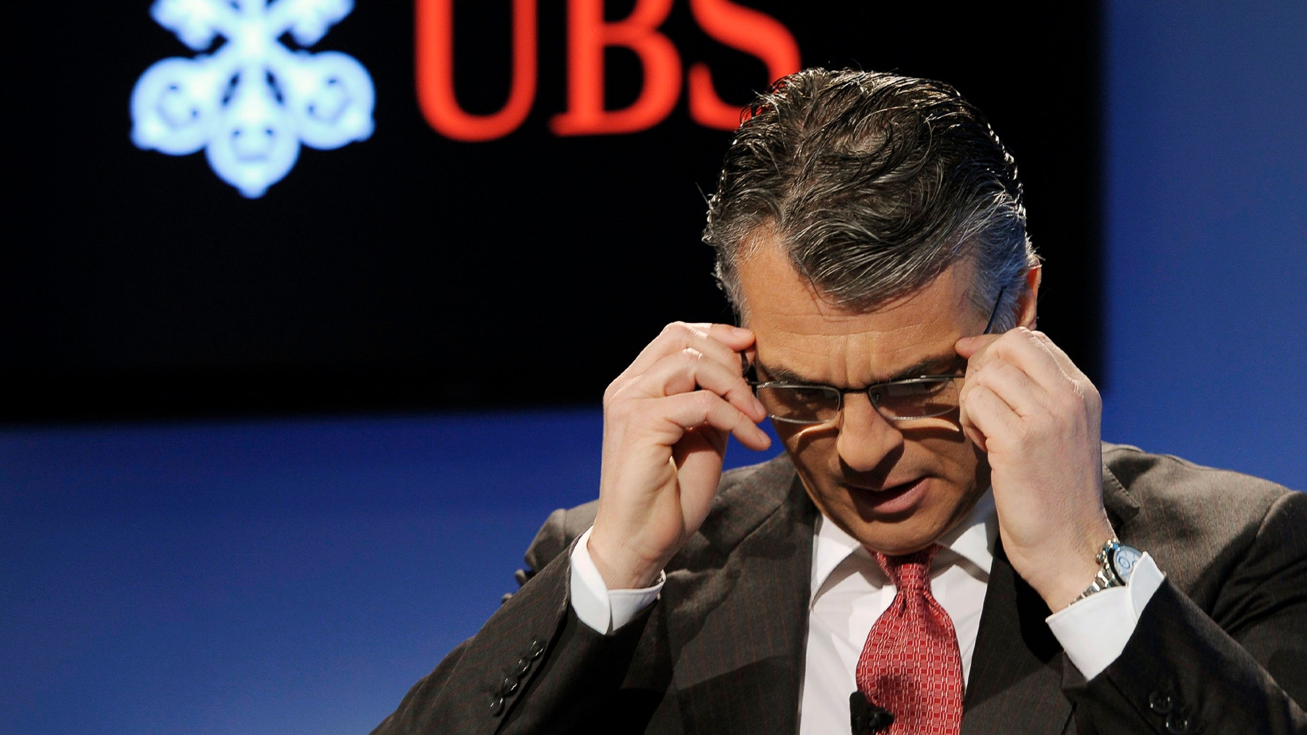 Man i mörk kostym och röd slips justerar sina glasögon på en scen med UBS-logotyp i bakgrunden.Sergio Ermotti, chef UBS.