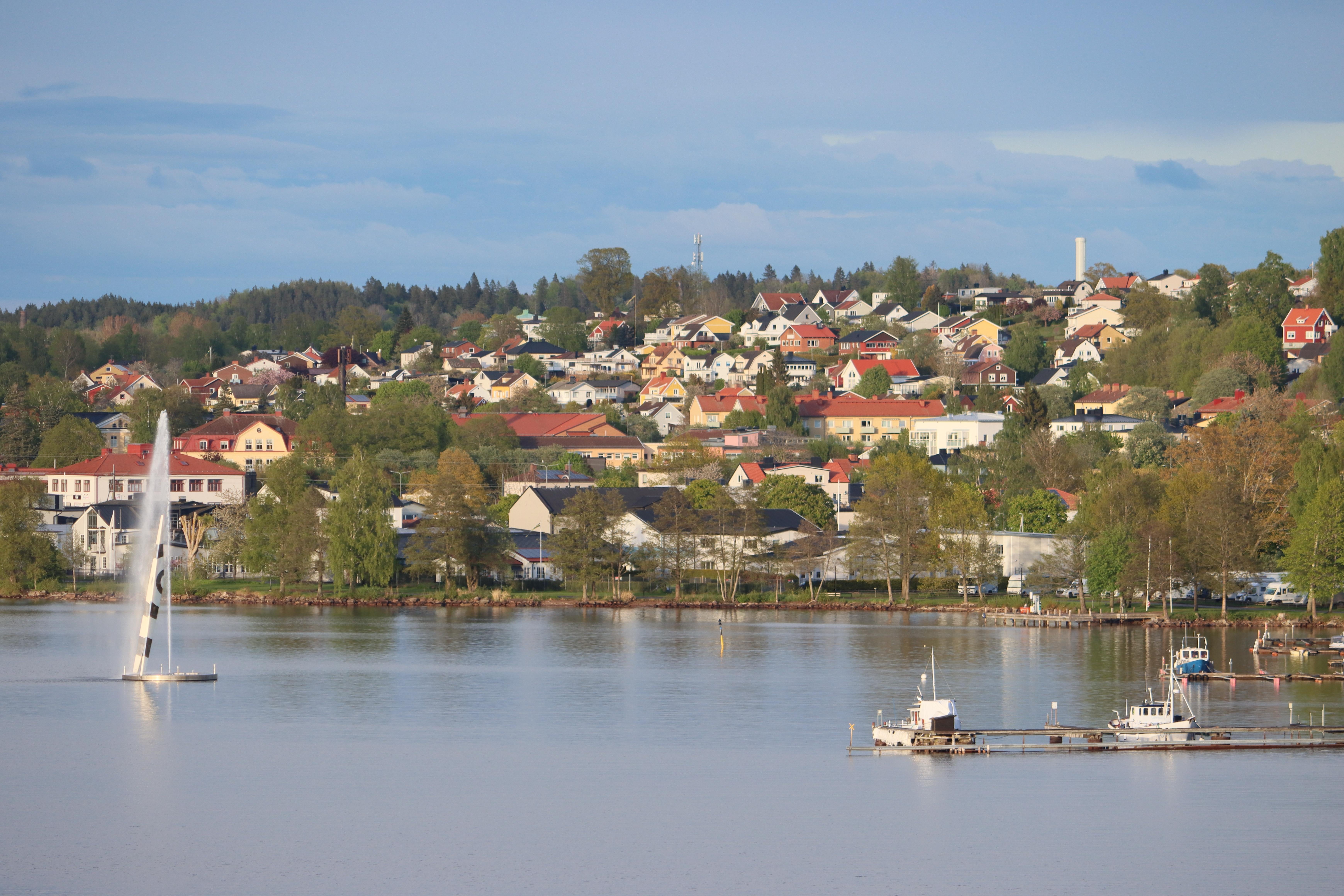 Utsikt över en svensk kuststad med villor på en sluttning vid vattnet. Färgglada hus, gröna träd och båtar syns i bilden.