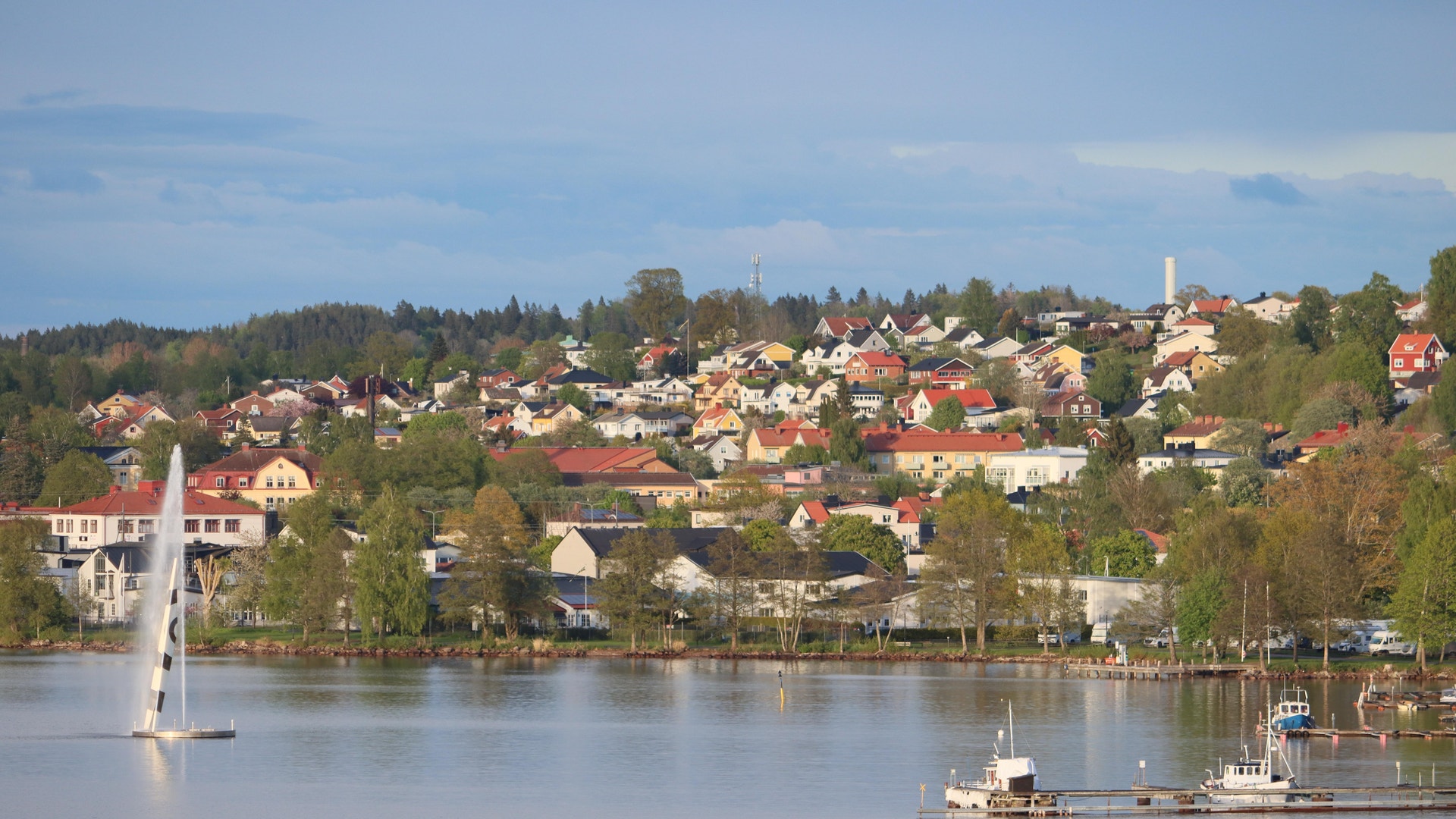 Utsikt över en svensk kuststad med villor på en sluttning vid vattnet. Färgglada hus, gröna träd och båtar syns i bilden.