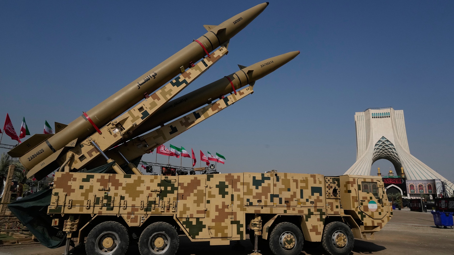 Två missiler på en militär lastbil utställda utomhus, med Azadi-tornet i Teheran i bakgrunden.