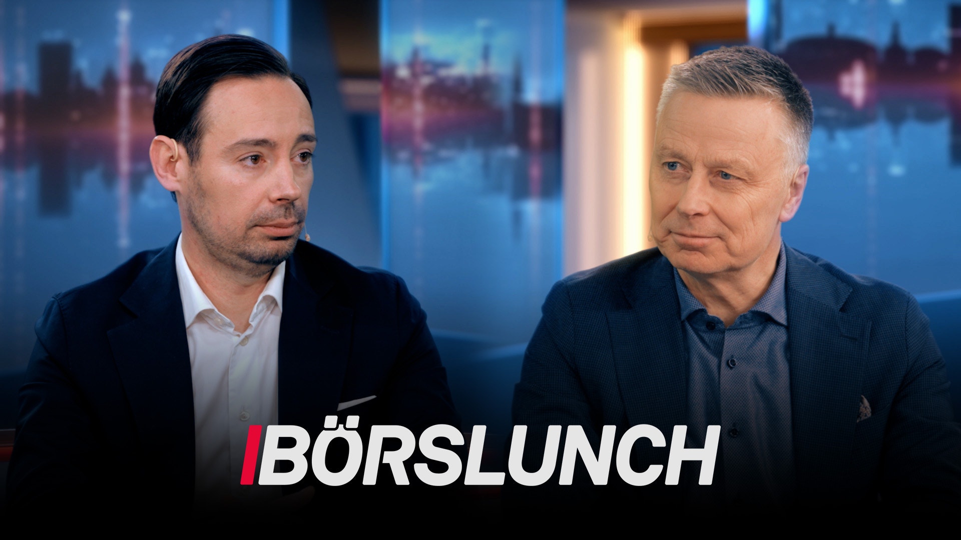 Två män i affärskläder diskuterar i en tv-studio med stadsutsikt i bakgrunden.