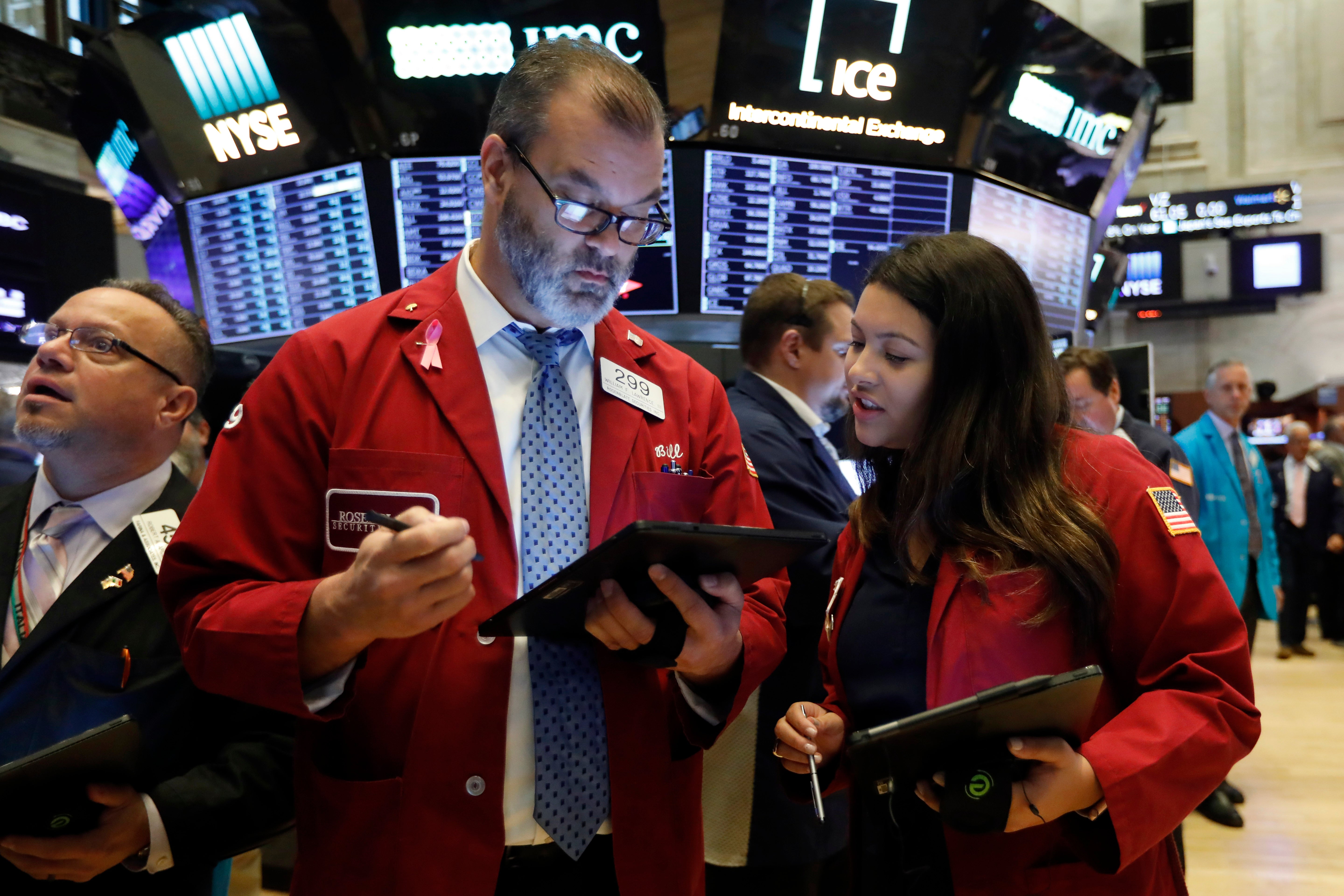 Handlare i röda jackor arbetar på börsgolvet med digitala enheter och övervakningsbildskärmar i bakgrunden.