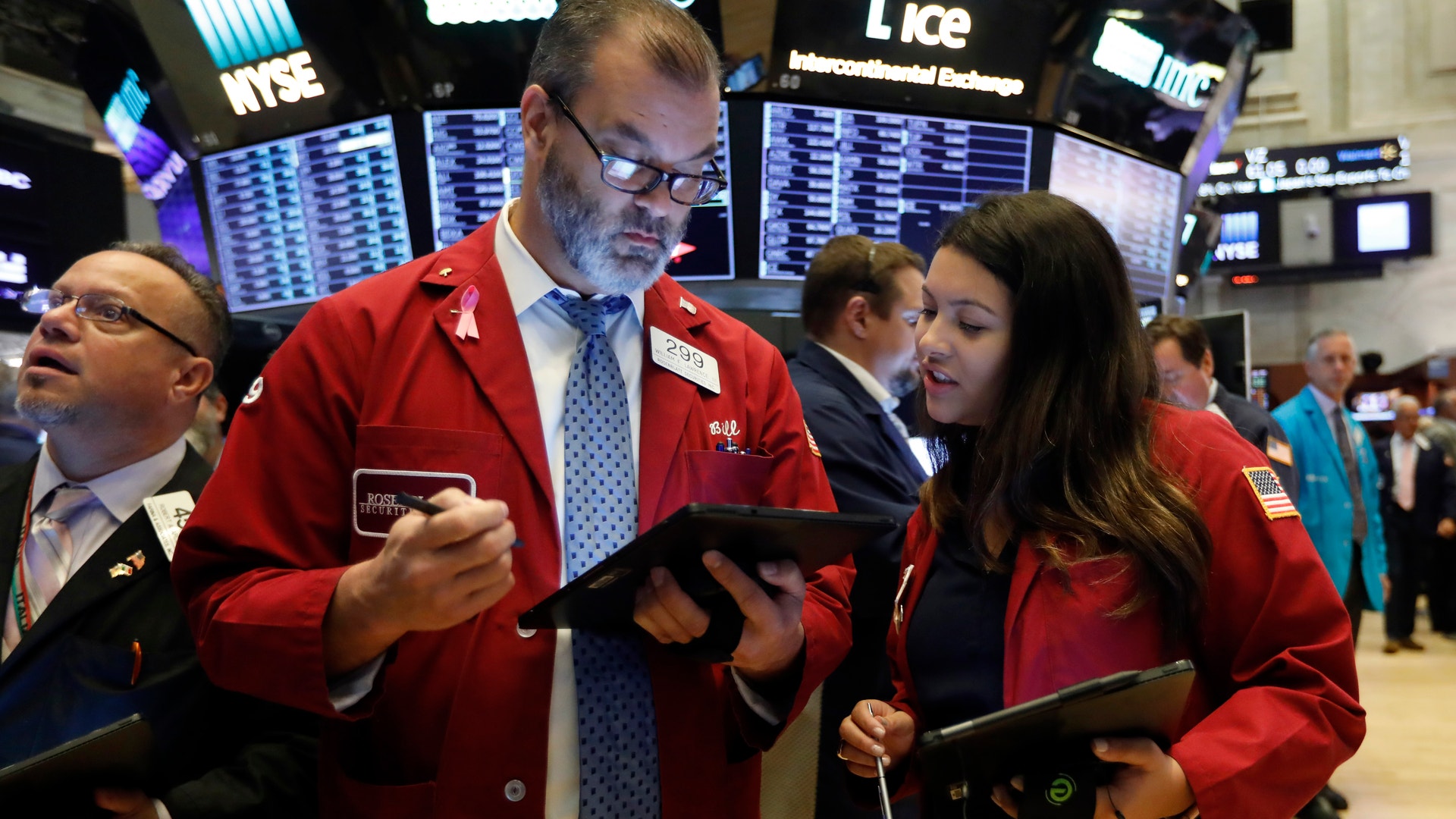 Handlare i röda jackor arbetar på börsgolvet med digitala enheter och övervakningsbildskärmar i bakgrunden.