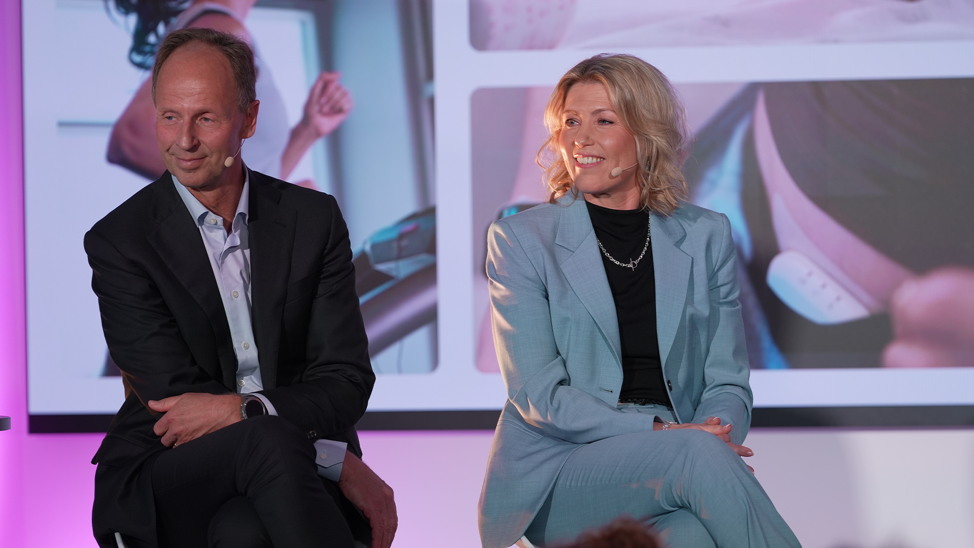 En man och en kvinna sitter på en scen under rosa belysning och deltar i en diskussion vid ett event.