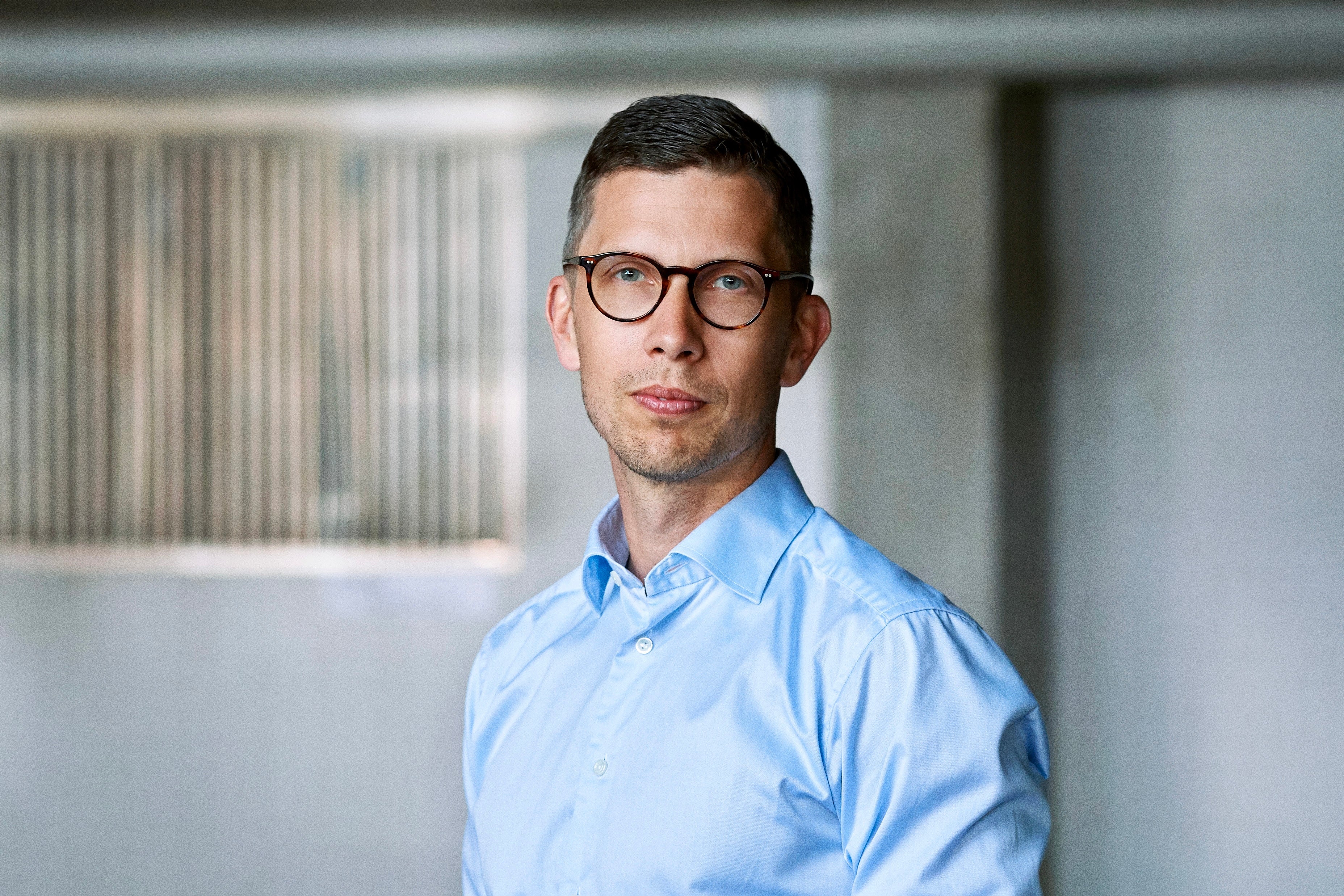 Porträttfoto av en man med glasögon och ljusblå skjorta mot en kontorsvägg.