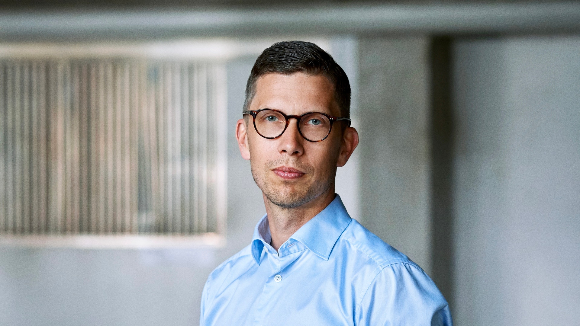 Porträttfoto av en man med glasögon och ljusblå skjorta mot en kontorsvägg.