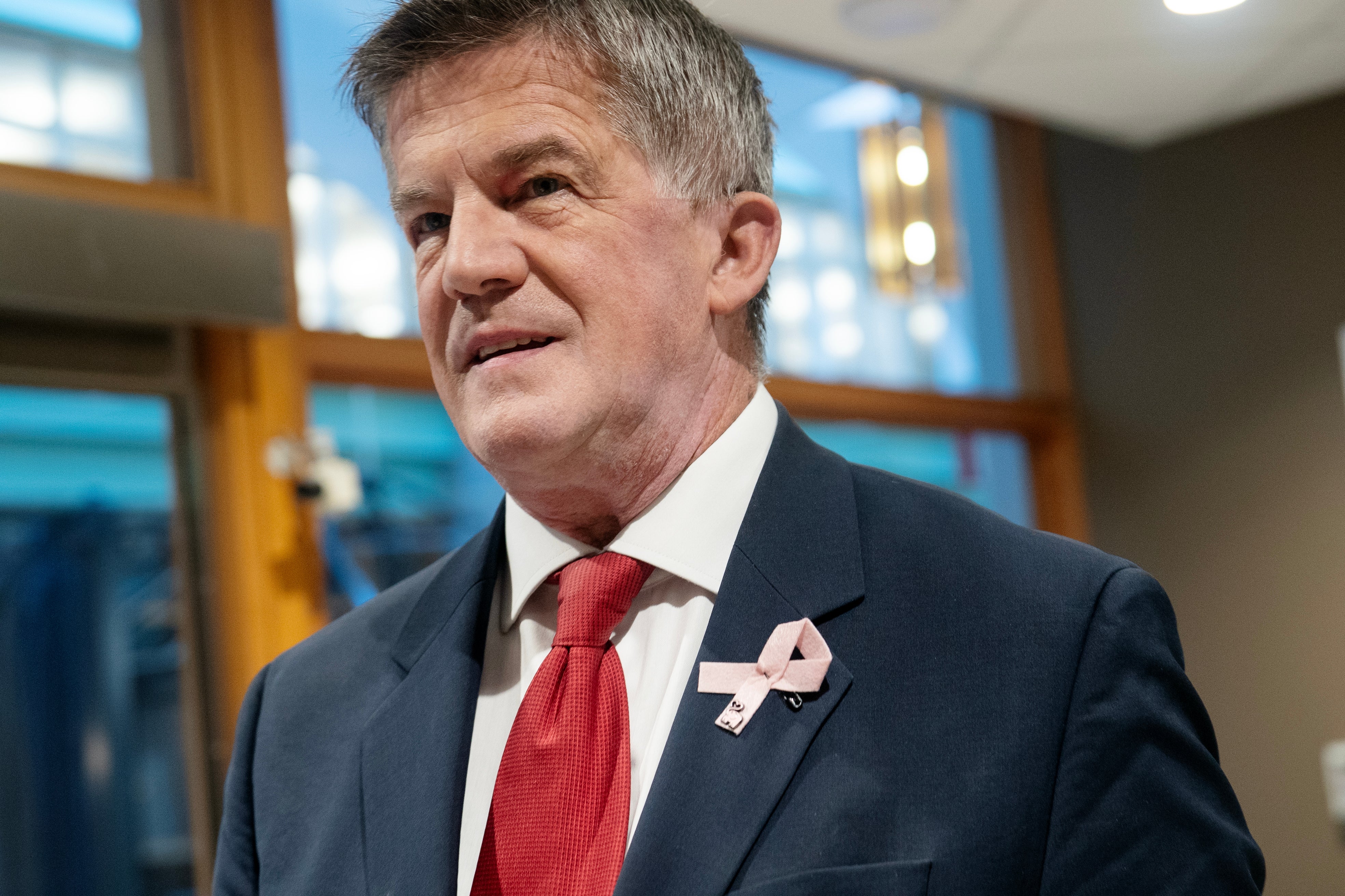 Man med grått hår i mörkblå kostym och röd slips, med ett rosa band på kavajslaget. Han tittar åt sidan och står inomhus.