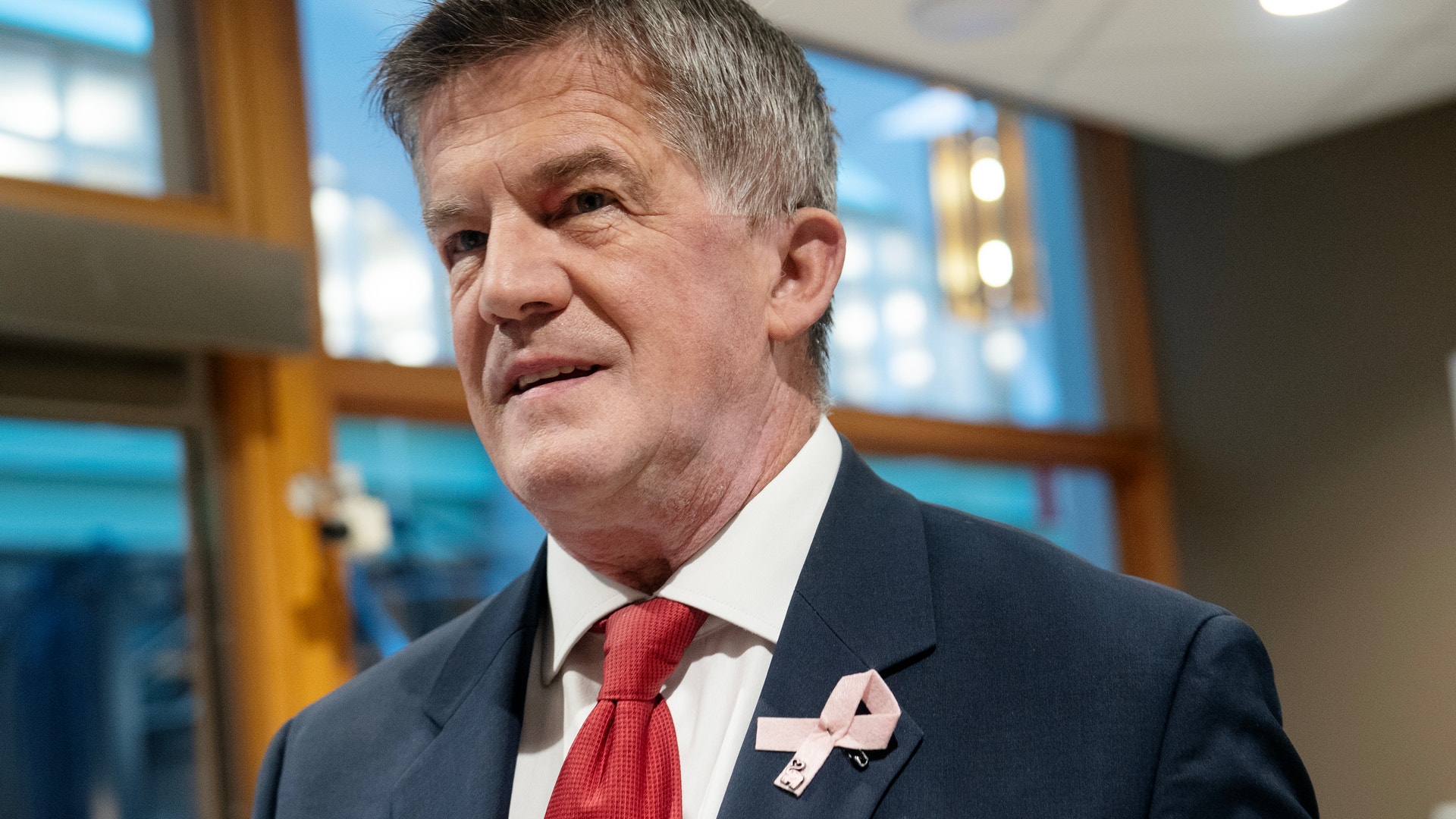 Man med grått hår i mörkblå kostym och röd slips, med ett rosa band på kavajslaget. Han tittar åt sidan och står inomhus.