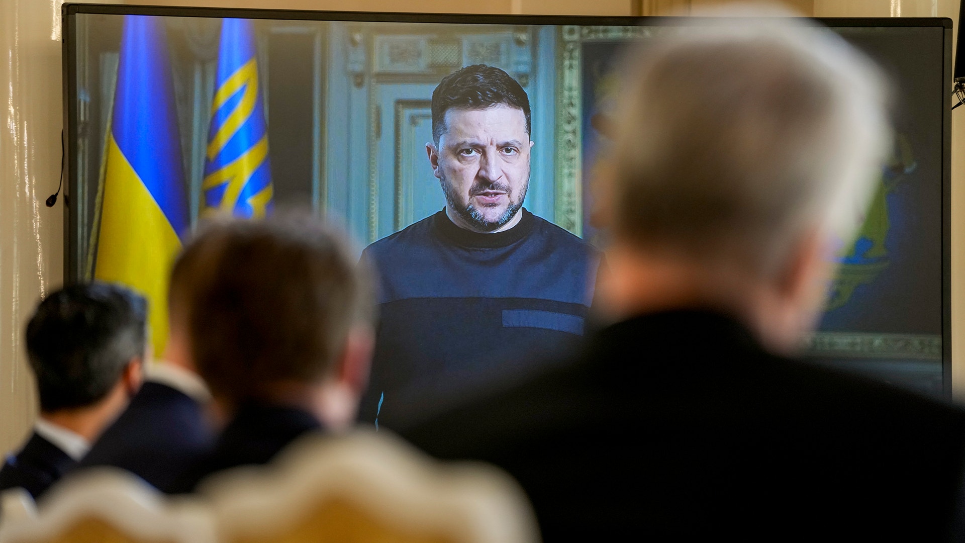 Man på videokonferens på skärm med ukrainska flaggor i bakgrunden, medan personer tittar på skärmen.