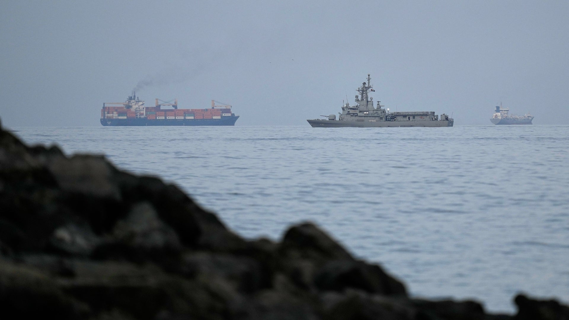 Flera fartyg på öppet hav, fotograferade från stranden med stenar i förgrunden.