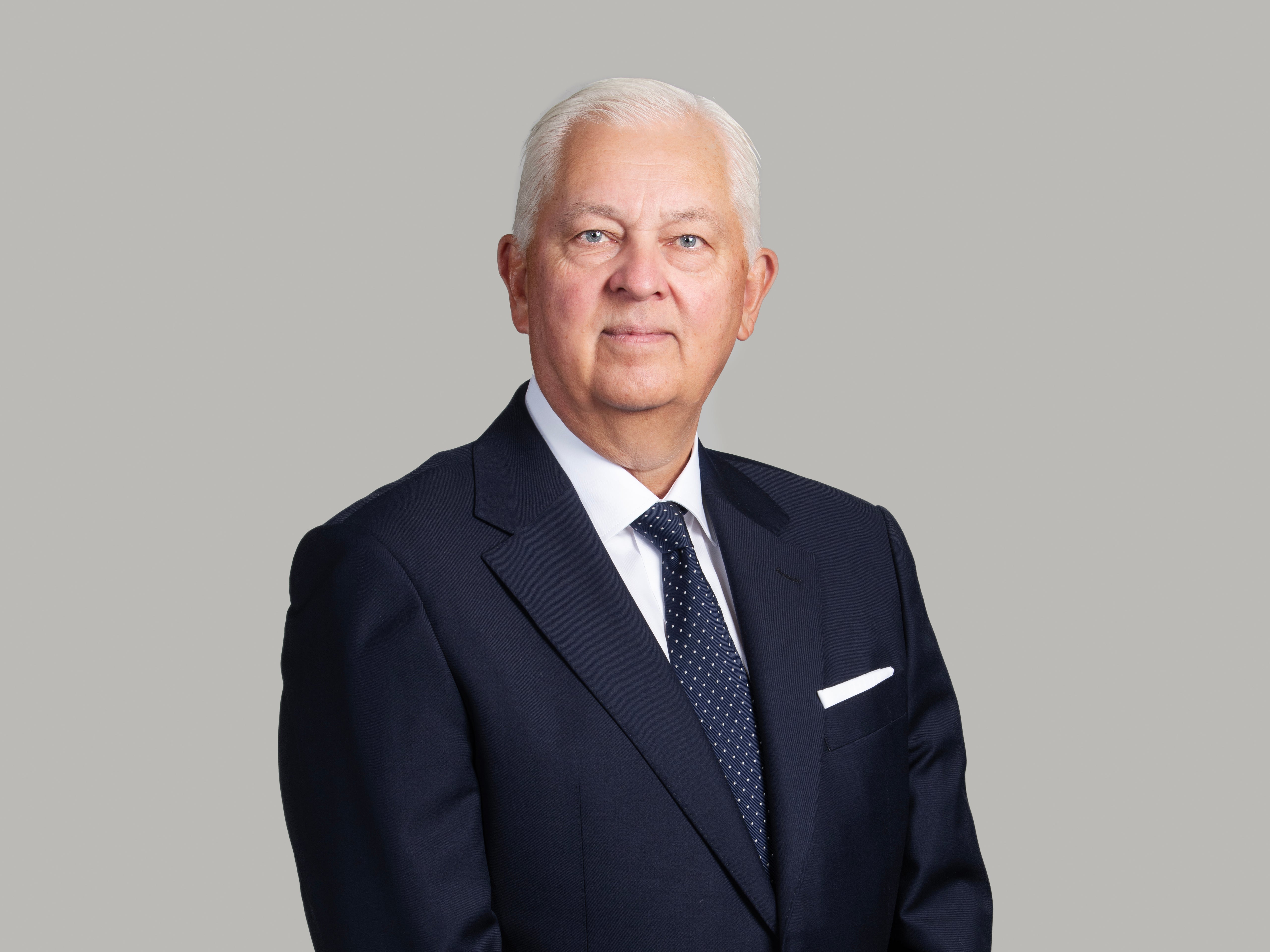 Porträttfoto av en äldre man i mörkblå kostym och slips mot grå bakgrund.