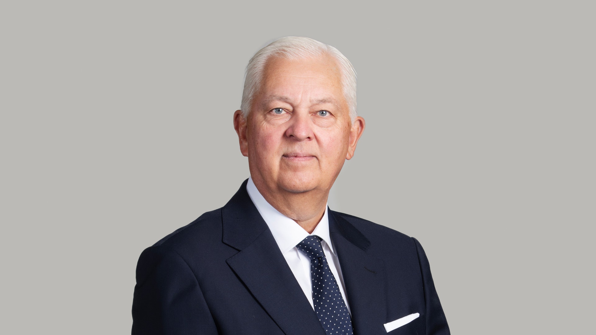 Porträttfoto av en äldre man i mörkblå kostym och slips mot grå bakgrund.