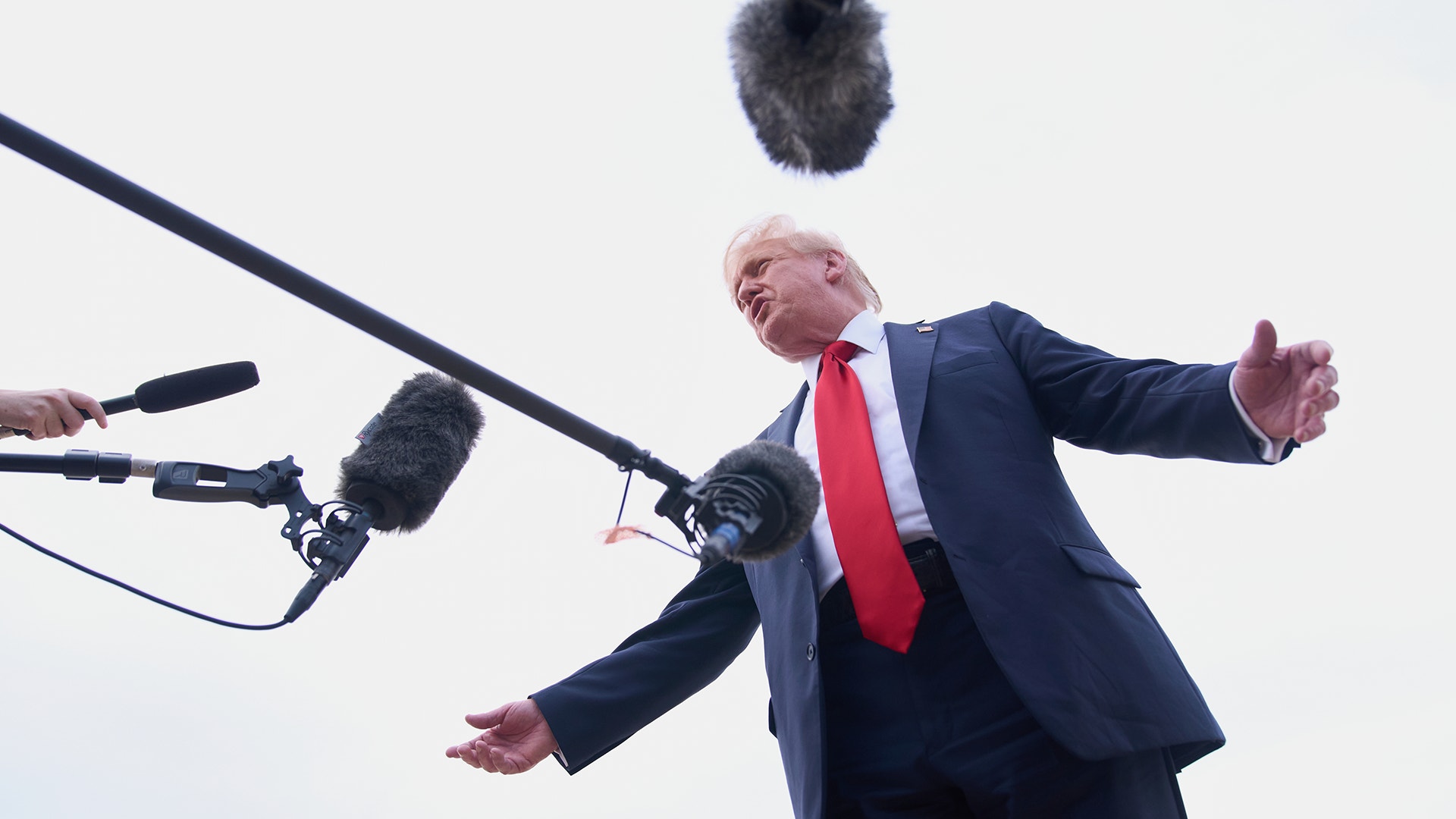 En bild på Donald Trump underifrån där han blir intervjuad med flera mikrofoner framför sig, i förgrunden av en vit, molnig himmel.