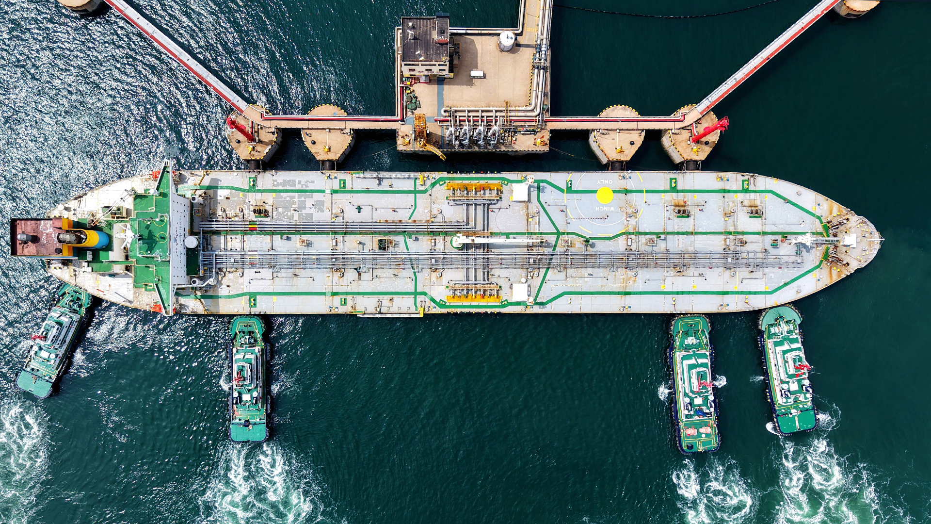 Flygfoto över ett stort tankfartyg dockat vid en kaj med rörsystem och försörjningsfartyg.