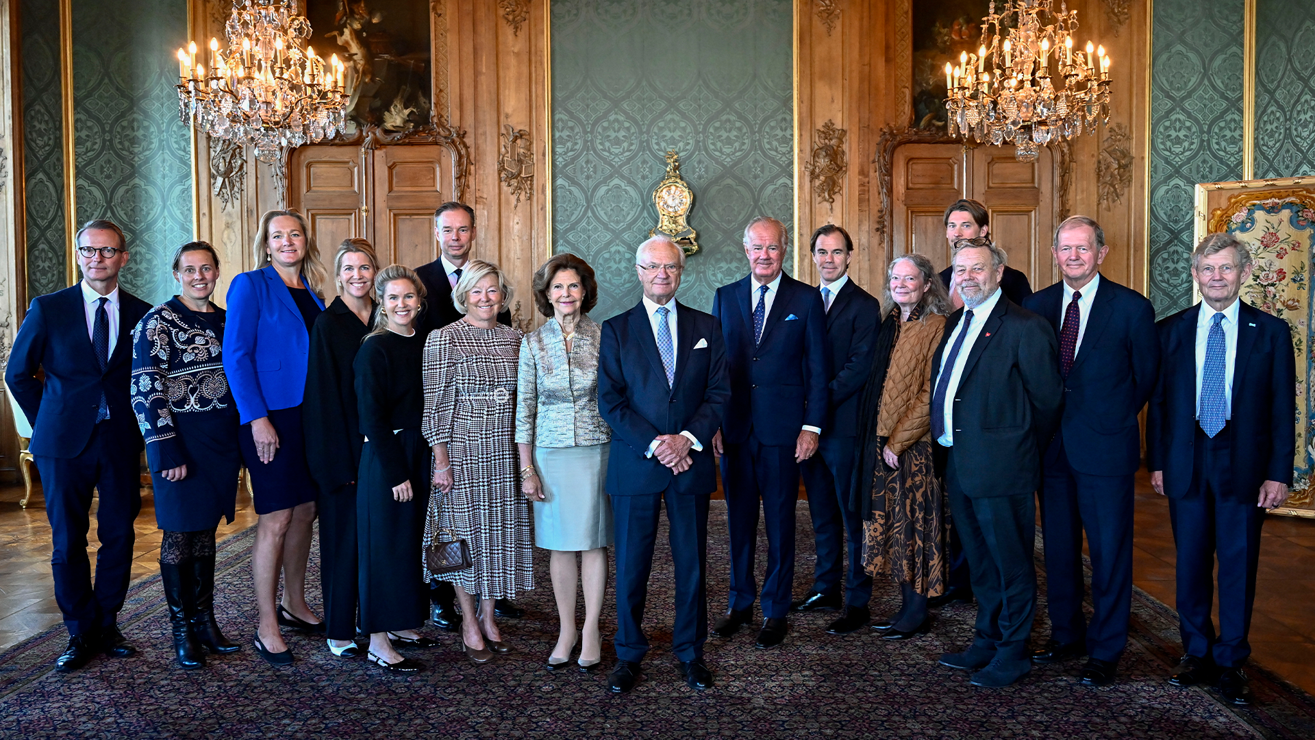Gruppfoto av omkring tjugo personer i formell klädsel som står tillsammans i ett elegant rum med kristallkronor och gulddekorationer.
