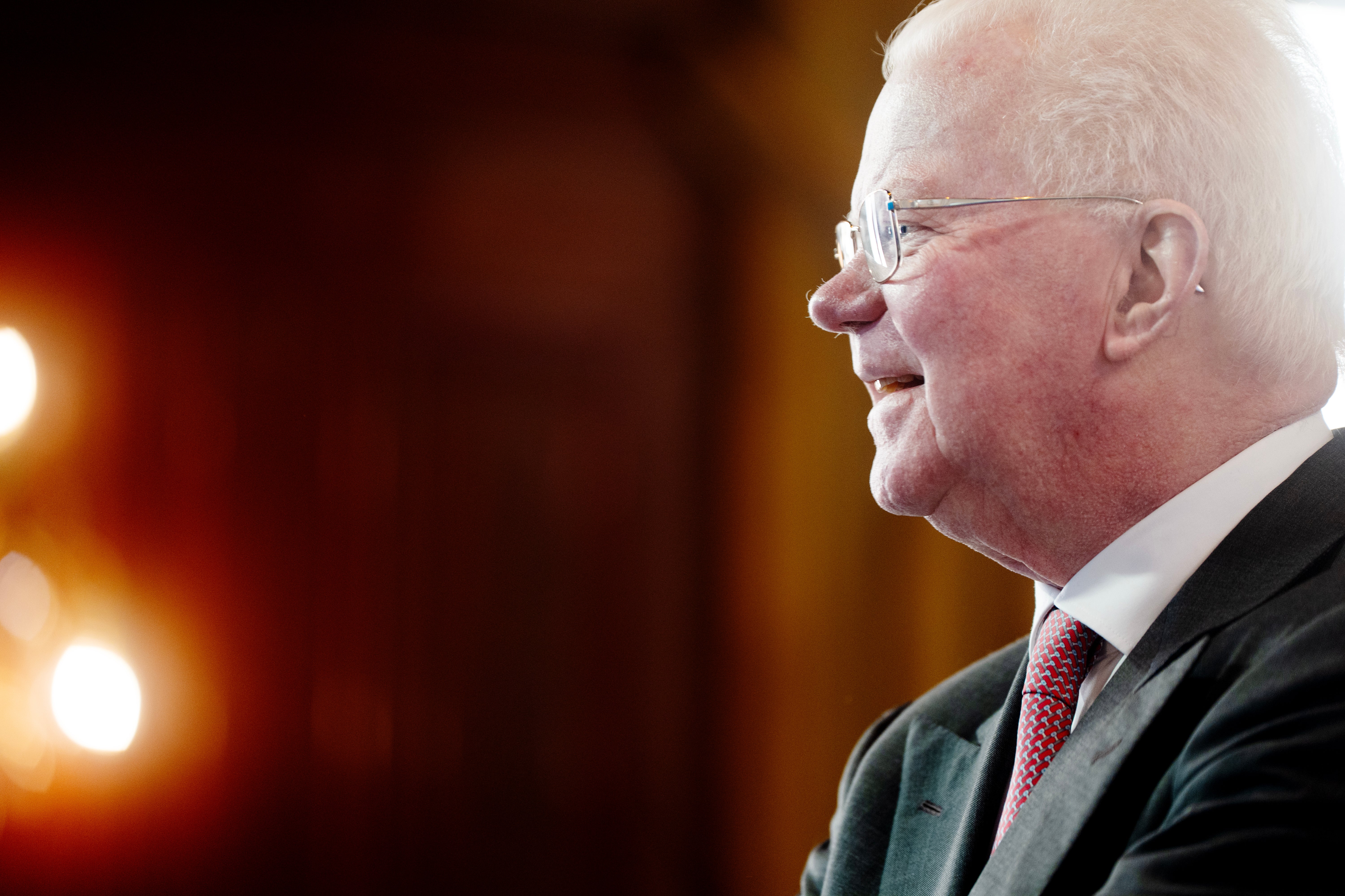 Äldre man i profil, iklädd mörk kostym och röd slips, mot en suddig bakgrund med varm belysning.