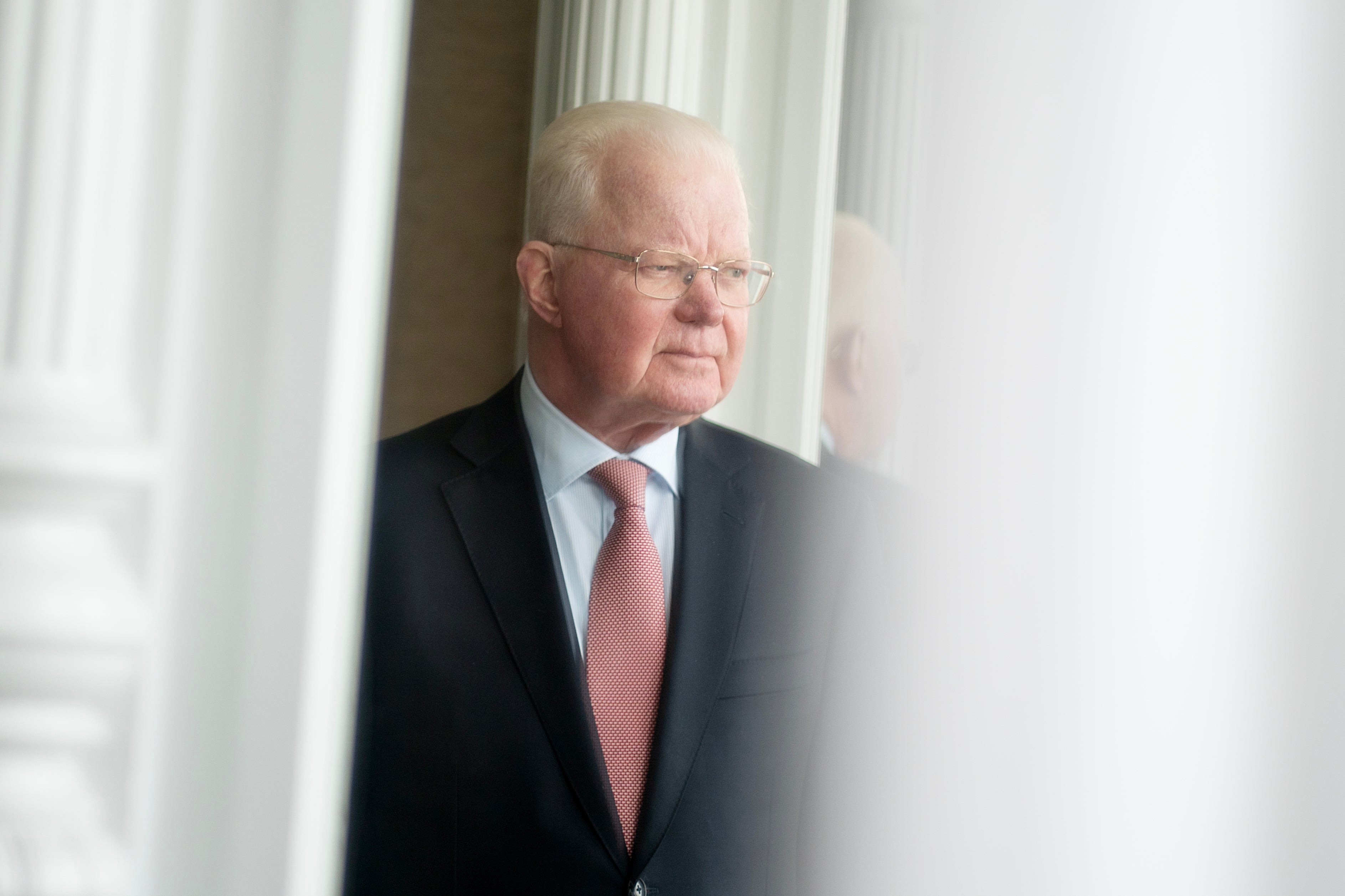 Porträtt av en äldre man i mörk kostym och röd slips framför ett fönster med vita gardiner.