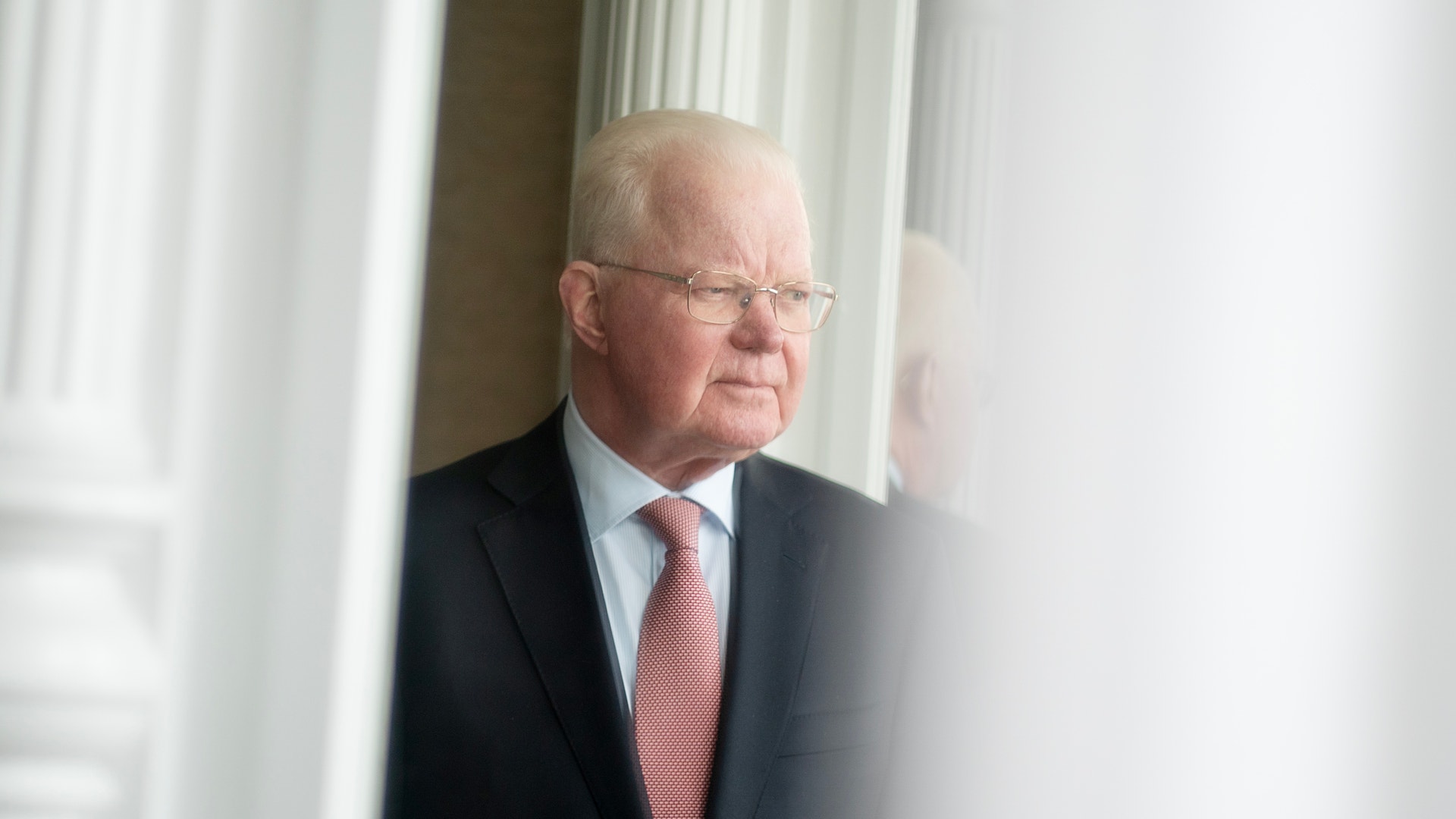 Porträtt av en äldre man i mörk kostym och röd slips framför ett fönster med vita gardiner.