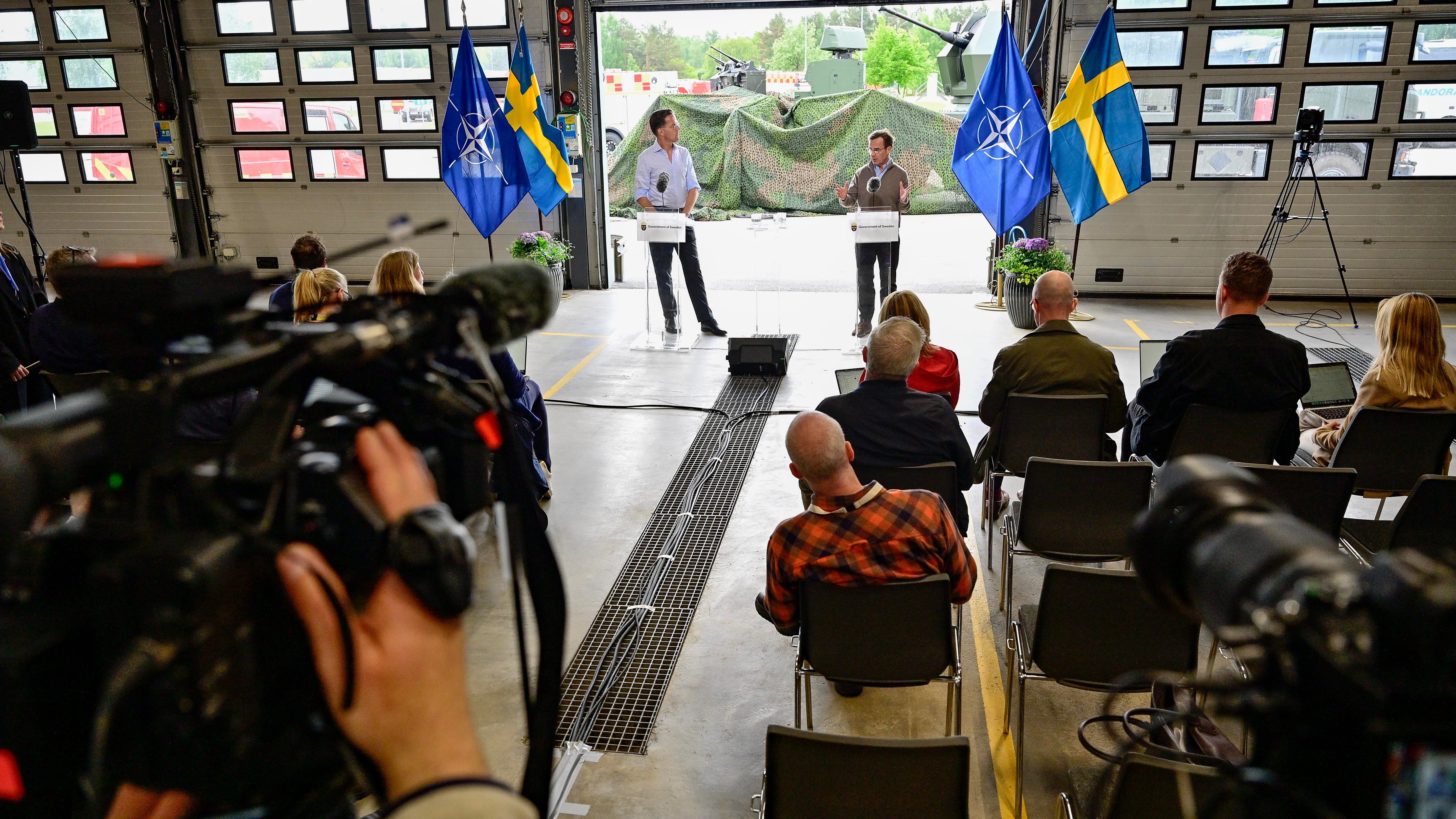En presskonferens eller offentligt evenemang med åskådare som sitter och tittar på två personer som står på en scen med svenska flaggor i bakgrunden.